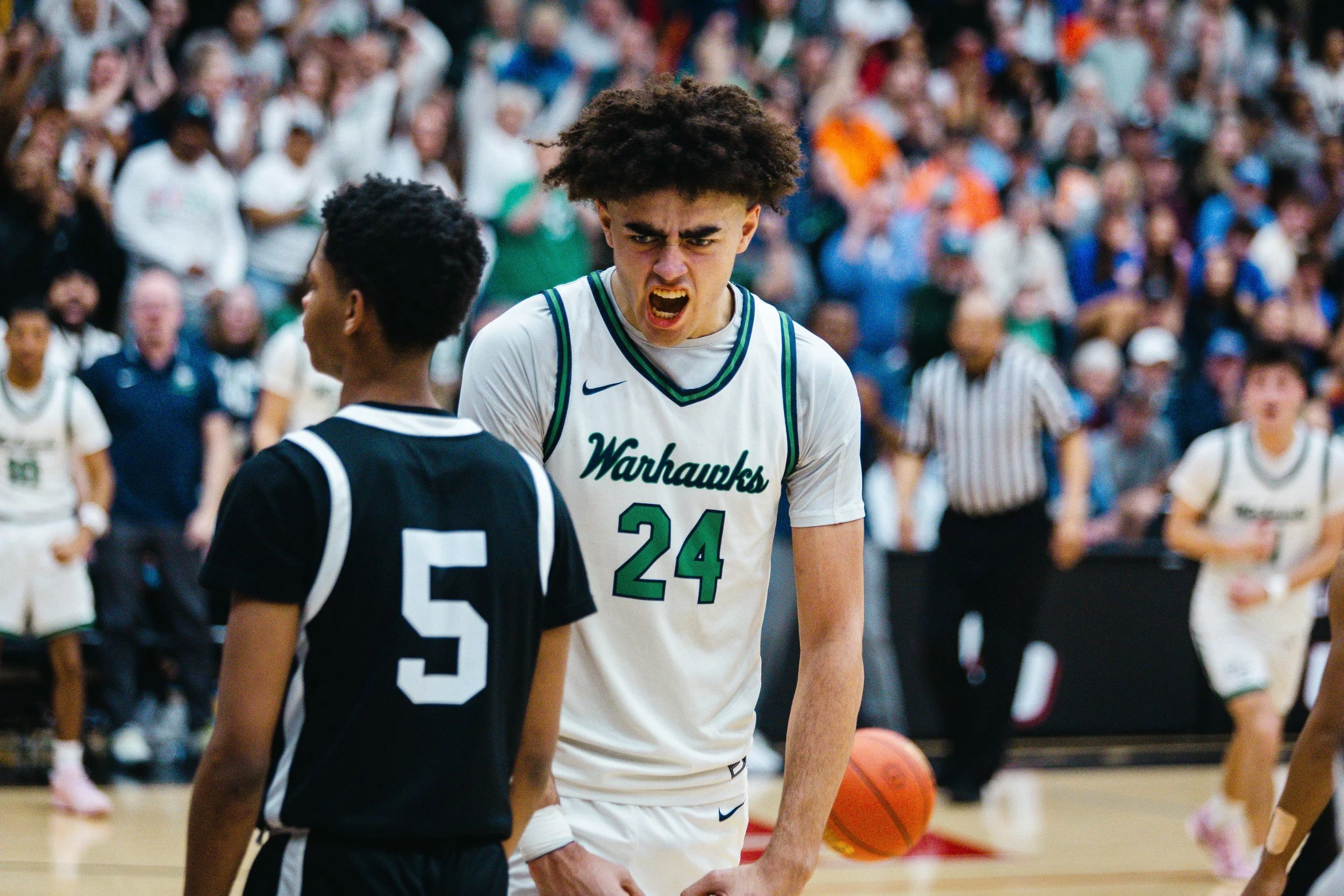 A basketball player wearing a white uniform with green accents and the number 24 from the Warhawks, appears angry or frustrated and is yelling at a referee or opponent during a game. There are other players and spectators in the background.
