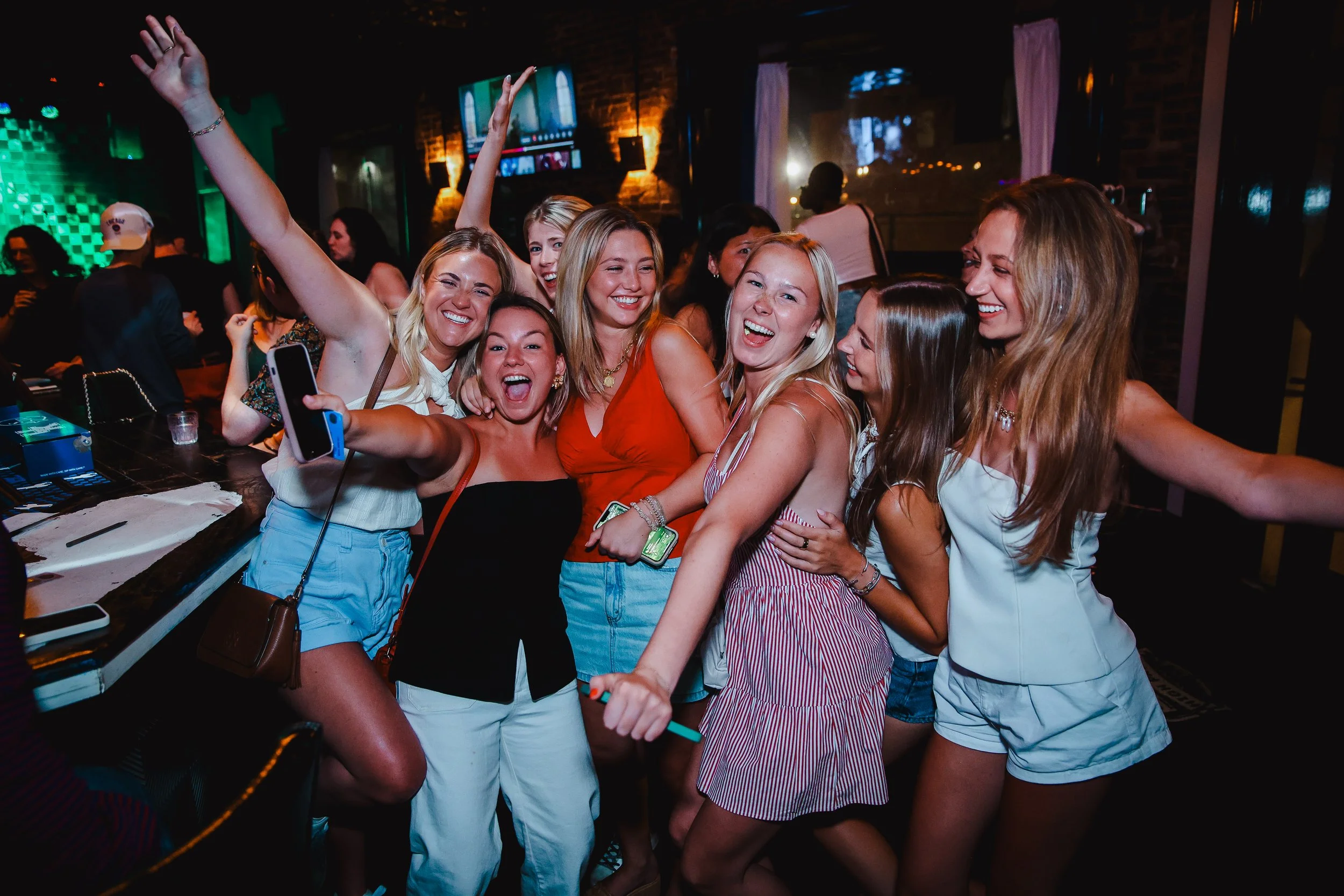Group of women joyfully posing and dancing in a bar or club setting with colorful lighting and televisions in the background.
