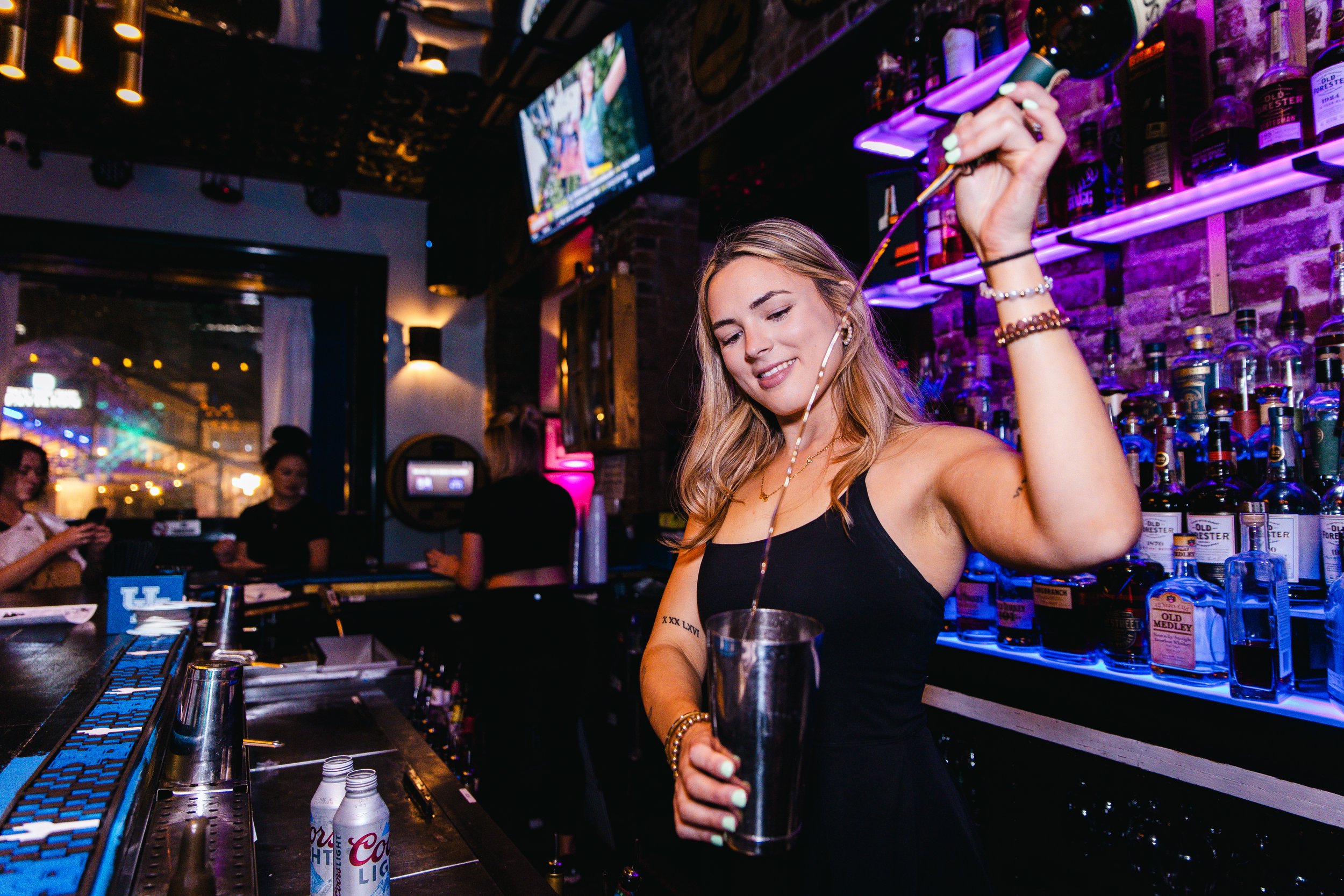 A young woman bartender with blonde hair, wearing a black tank top, is smiling and preparing a drink behind a bar. The bar has various liquor bottles on illuminated shelves, and a TV screen is visible in the background with two other women and a pers