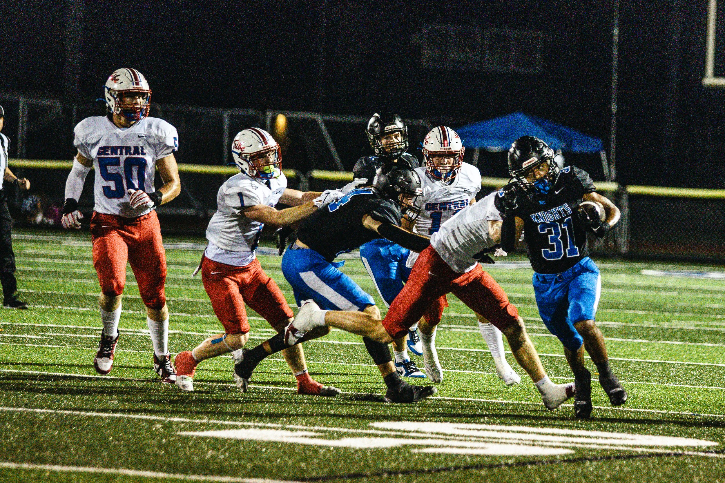 A nighttime football game with players from two teams, one in black and the other in white and red uniforms, in action on the field. The player in black is running with the ball, while players in white and red attempt to tackle him.