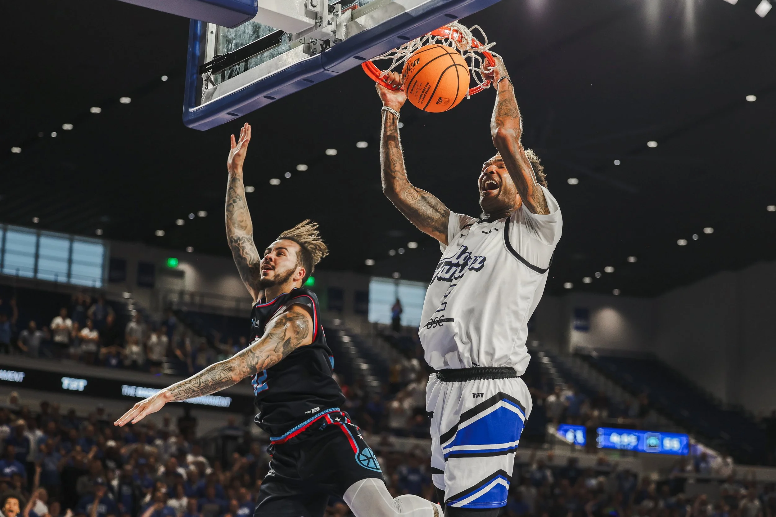 A basketball player in a white uniform is dunking the ball into the hoop while an opposing player in a black uniform attempts to block the shot during a game.