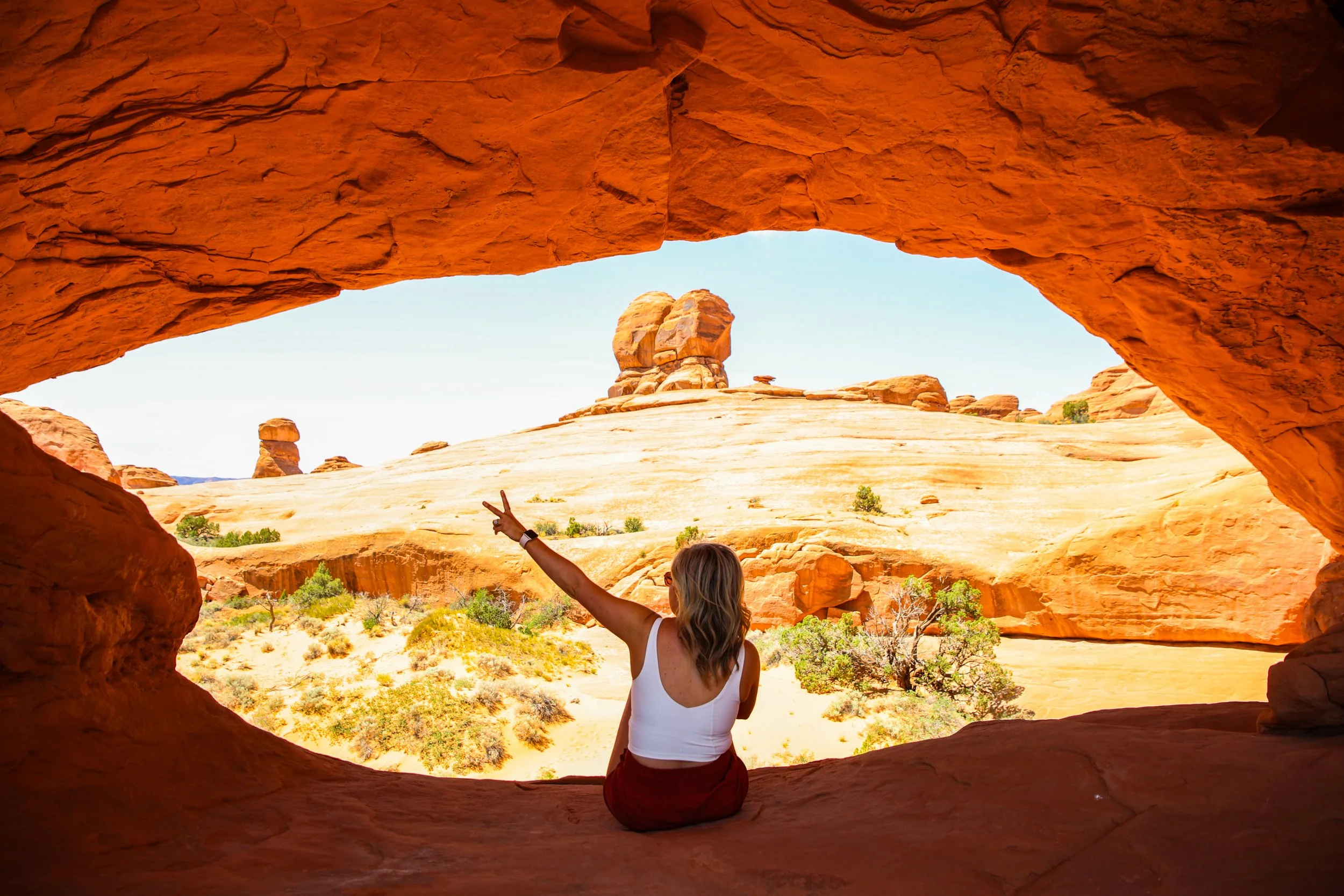 A woman sitting in a natural rock formation view, making a peace sign with her hand, overlooking a desert landscape with large rock formations.