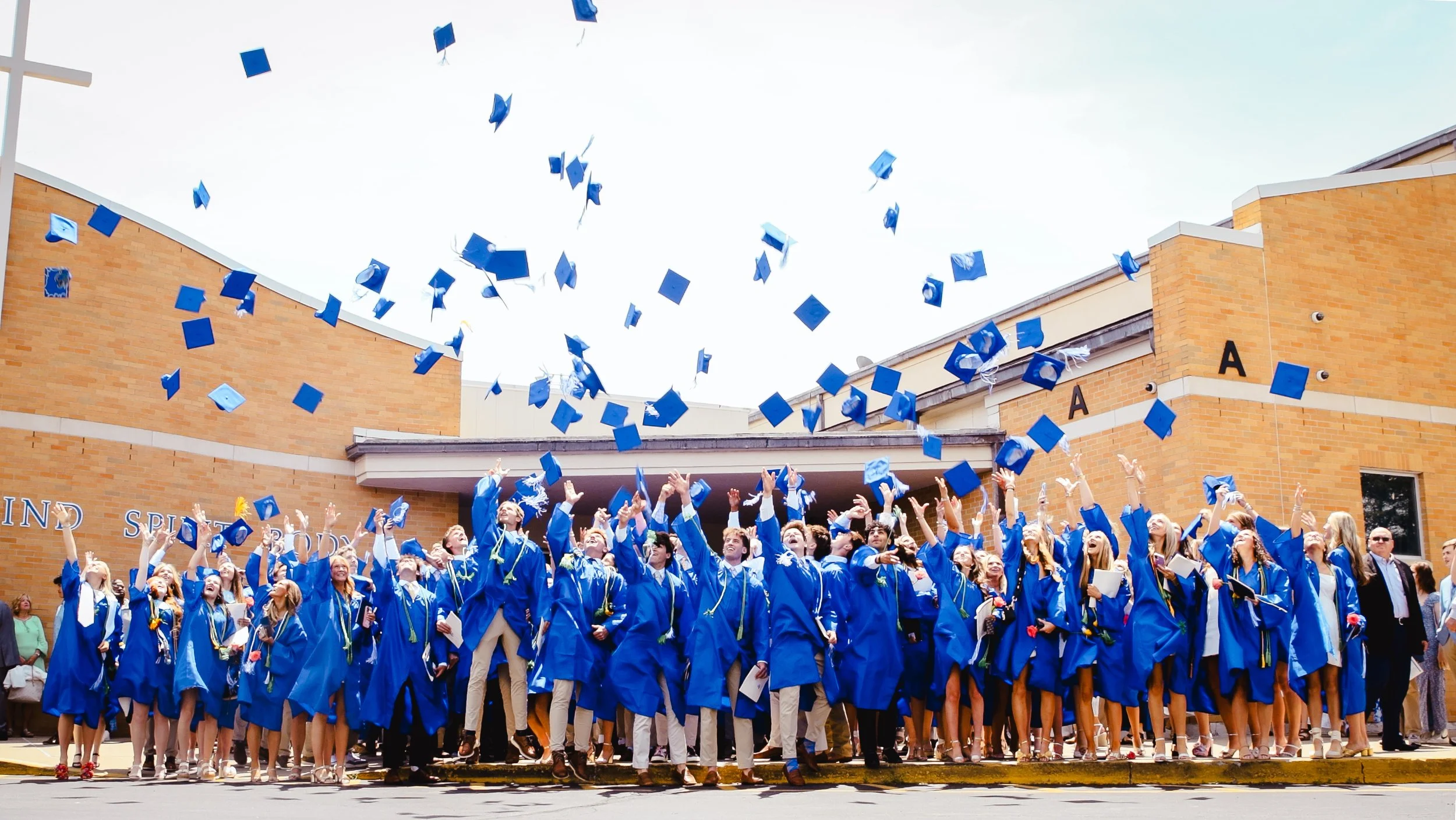 Graduates in blue caps and gowns celebrating outside a school building, throwing caps into the air during graduation ceremony.