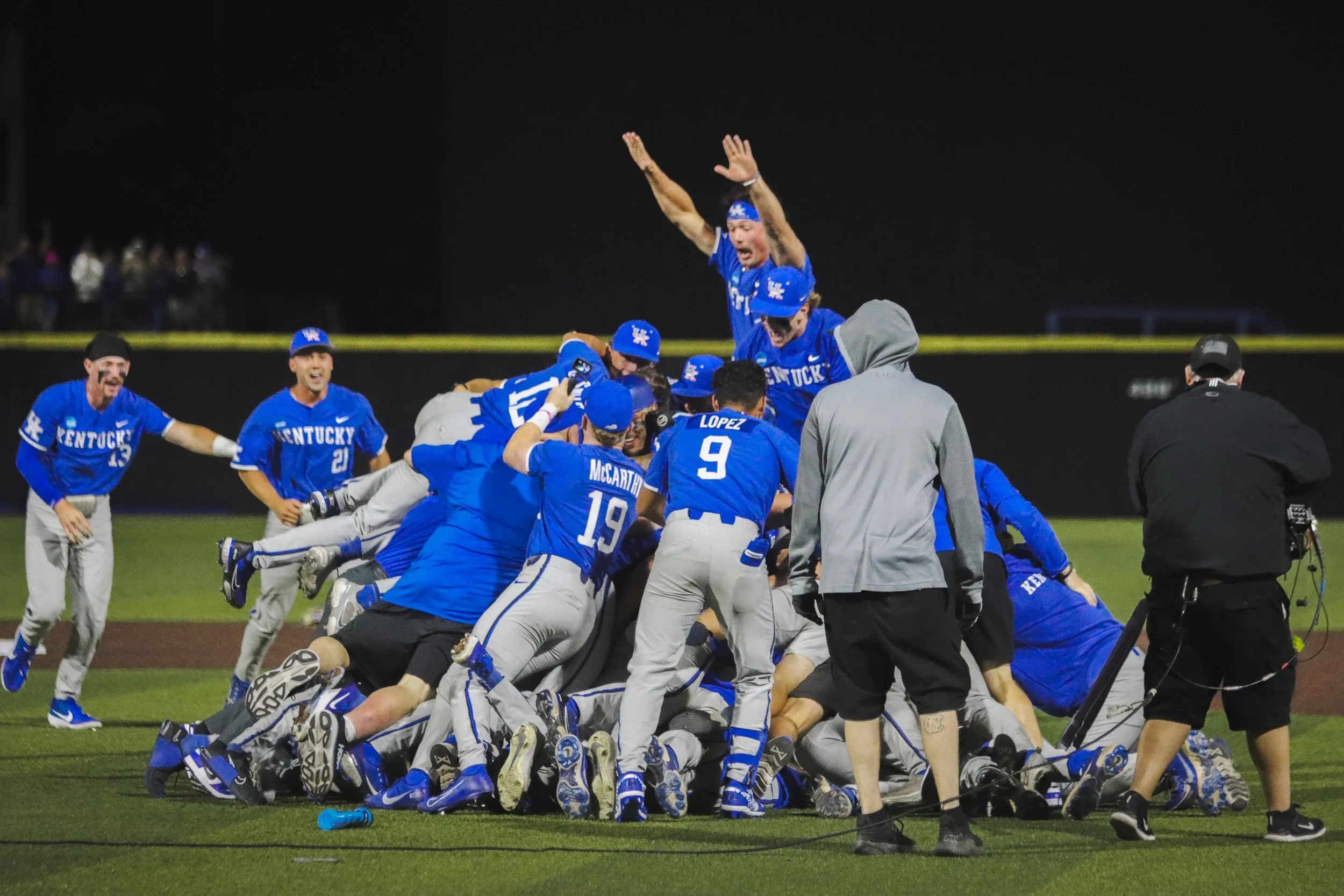Kentucky aseball players in blue uniforms celebrating a victory on the field at night with some players piled on top of each other and others standing around.