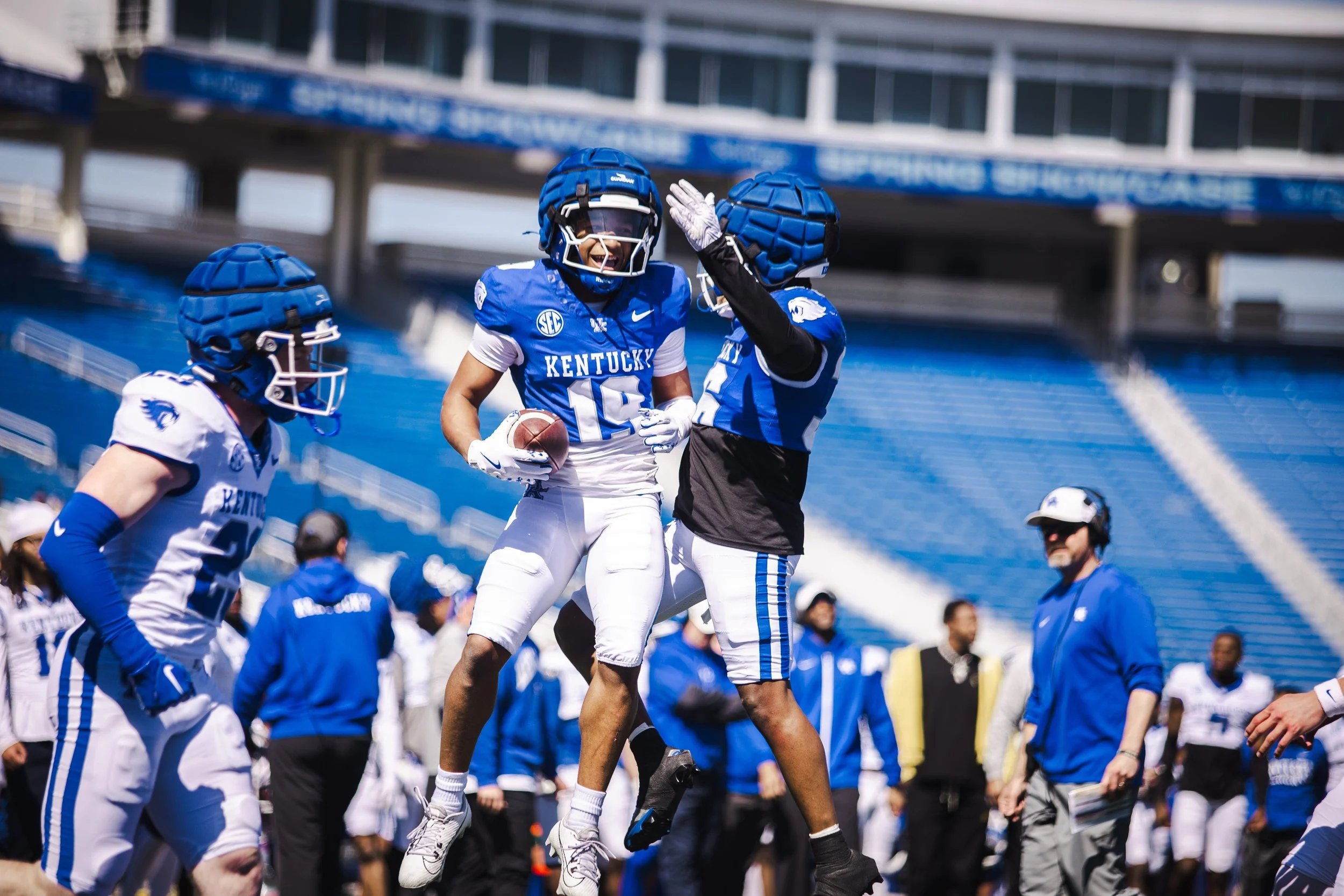 College football players from Kentucky celebrating on the field, one holding a football, with coaching staff and spectators in the background.