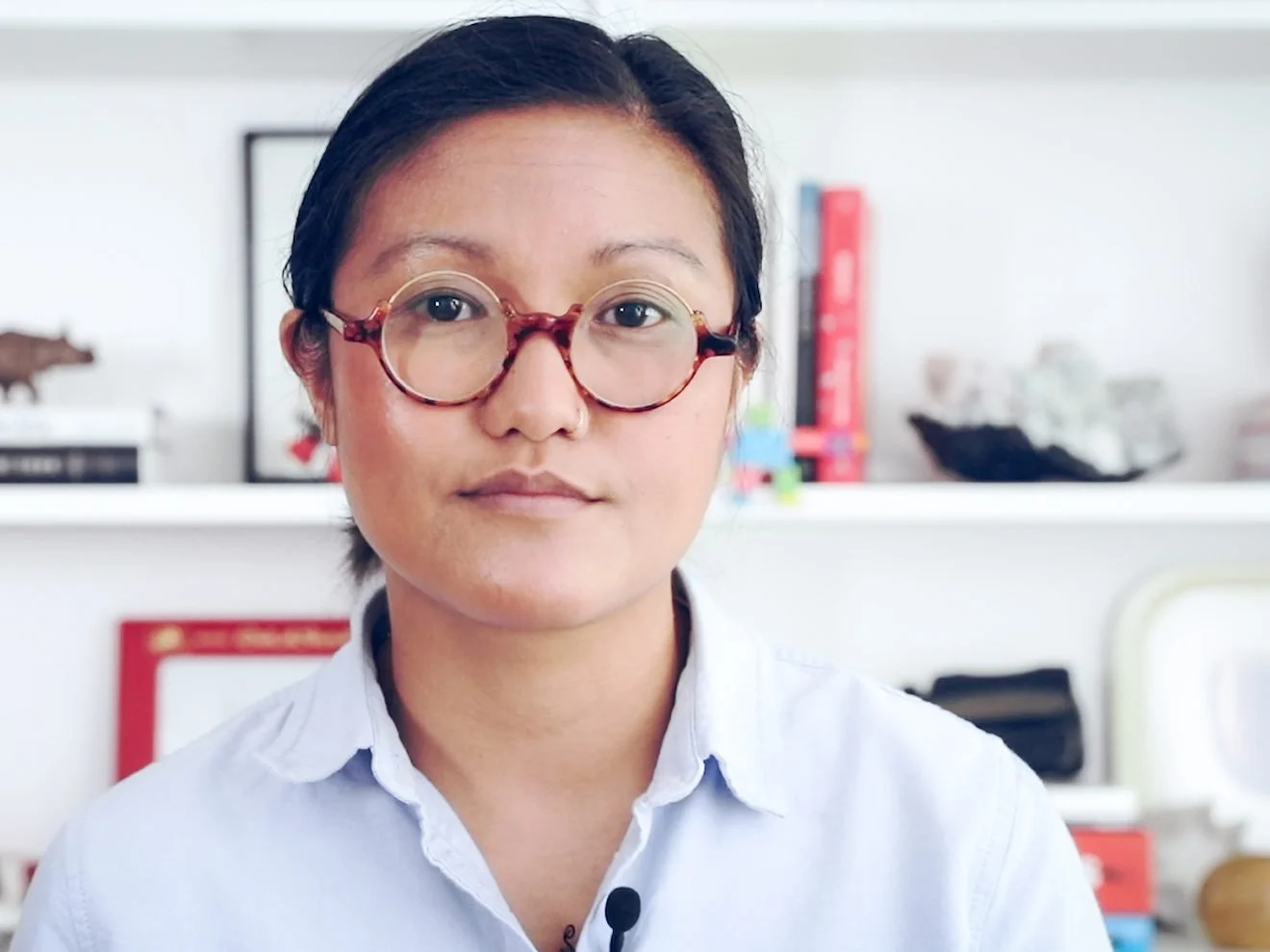 A medium close headshot of Paco standing in front of a white wall shelf that’s decorated with various books and paintings. Paco wears a light blue polo, round light brown glasses, and her hair pulled back.