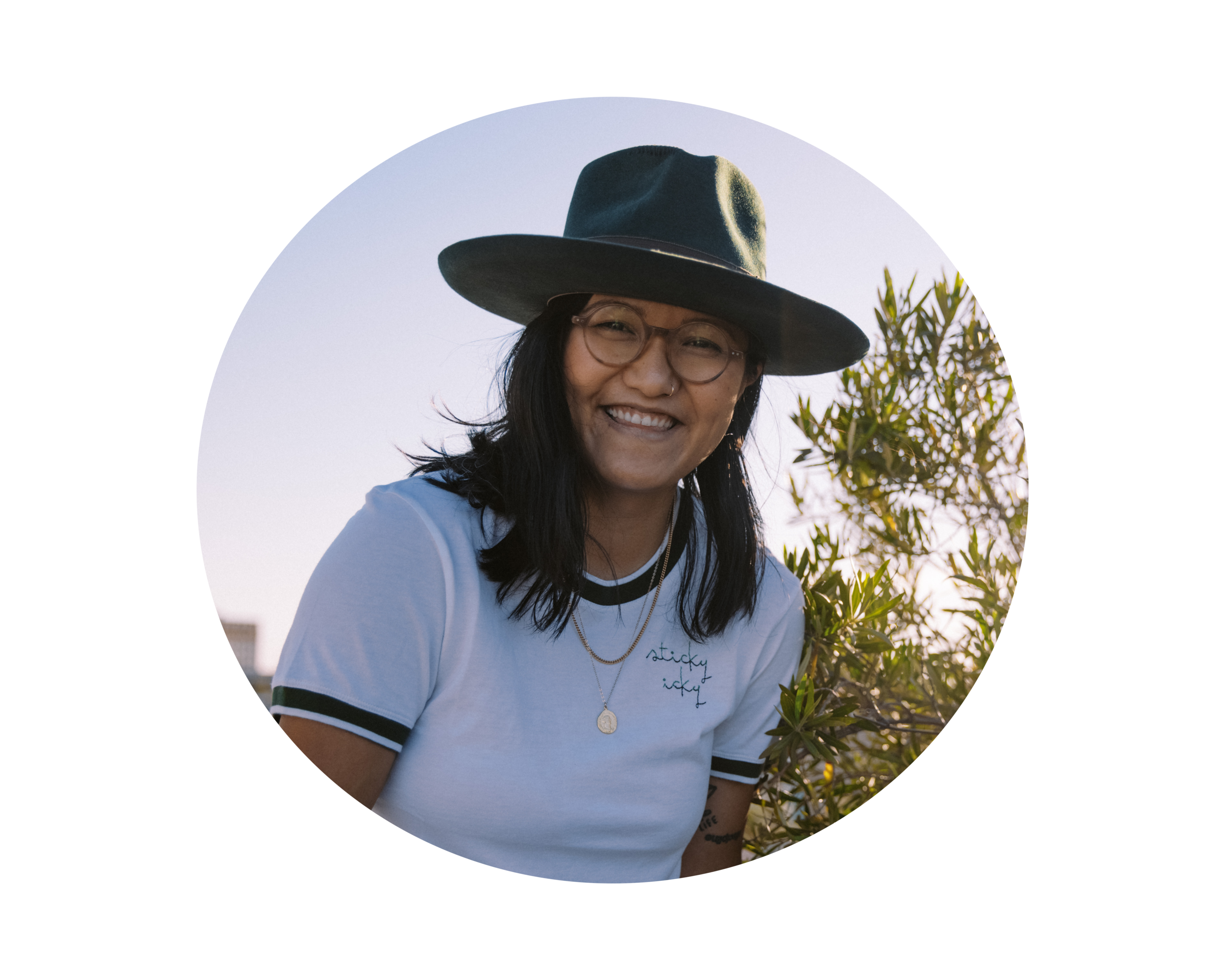 Standing in front of a leafy green tree in the daylight, Paco De Leon smiles mid-laughter. She wears a white t-shirt and a wide-brimmed olive green fedora. Her hair is down and she wears round glasses.