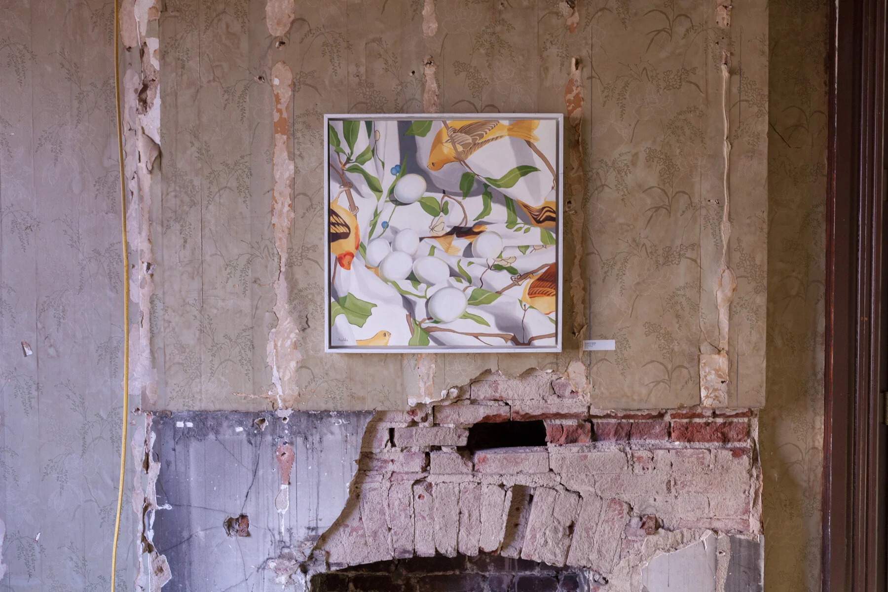 A painting depicting a branch with green leaves and yellow-orange fish or birds on a fabric surface, hung on a distressed wall with peeling wallpaper and exposed brickwork.