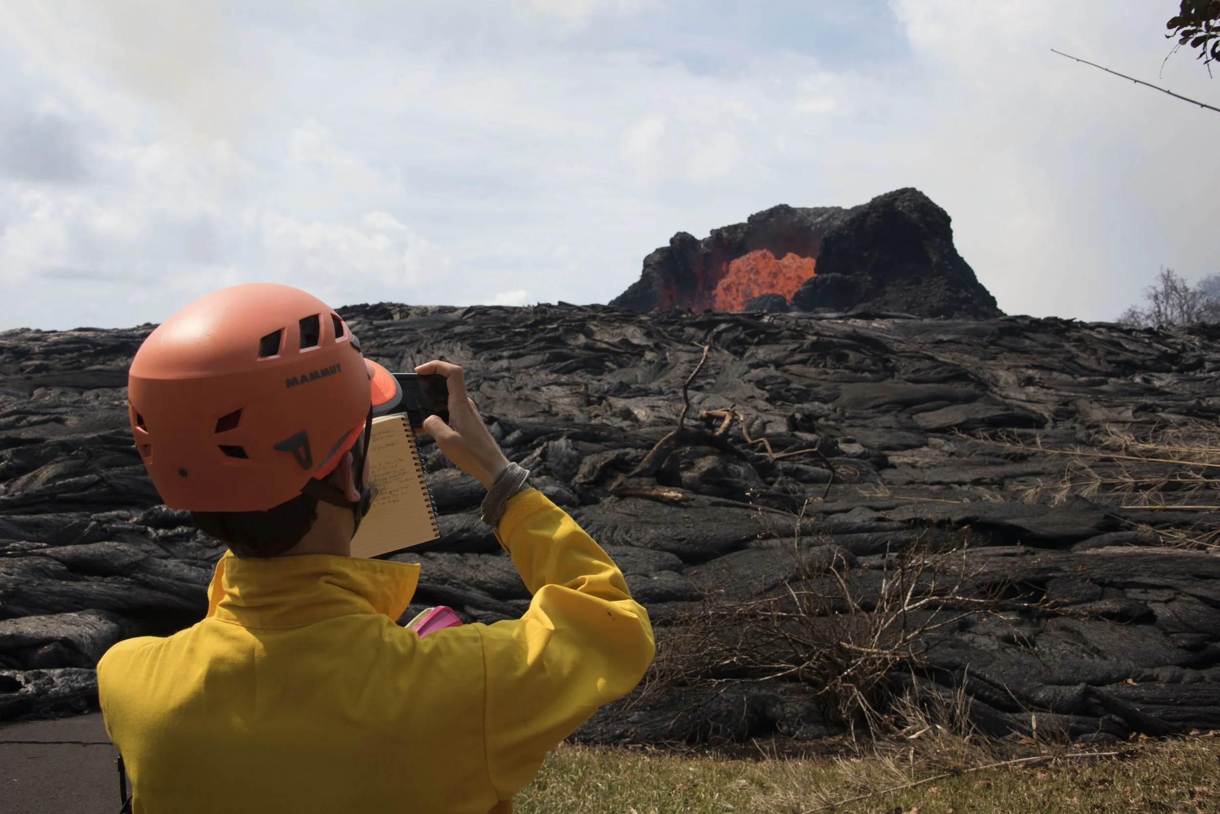 A volcanologist photographing an orange lava fountain amid dark volcanic rock.