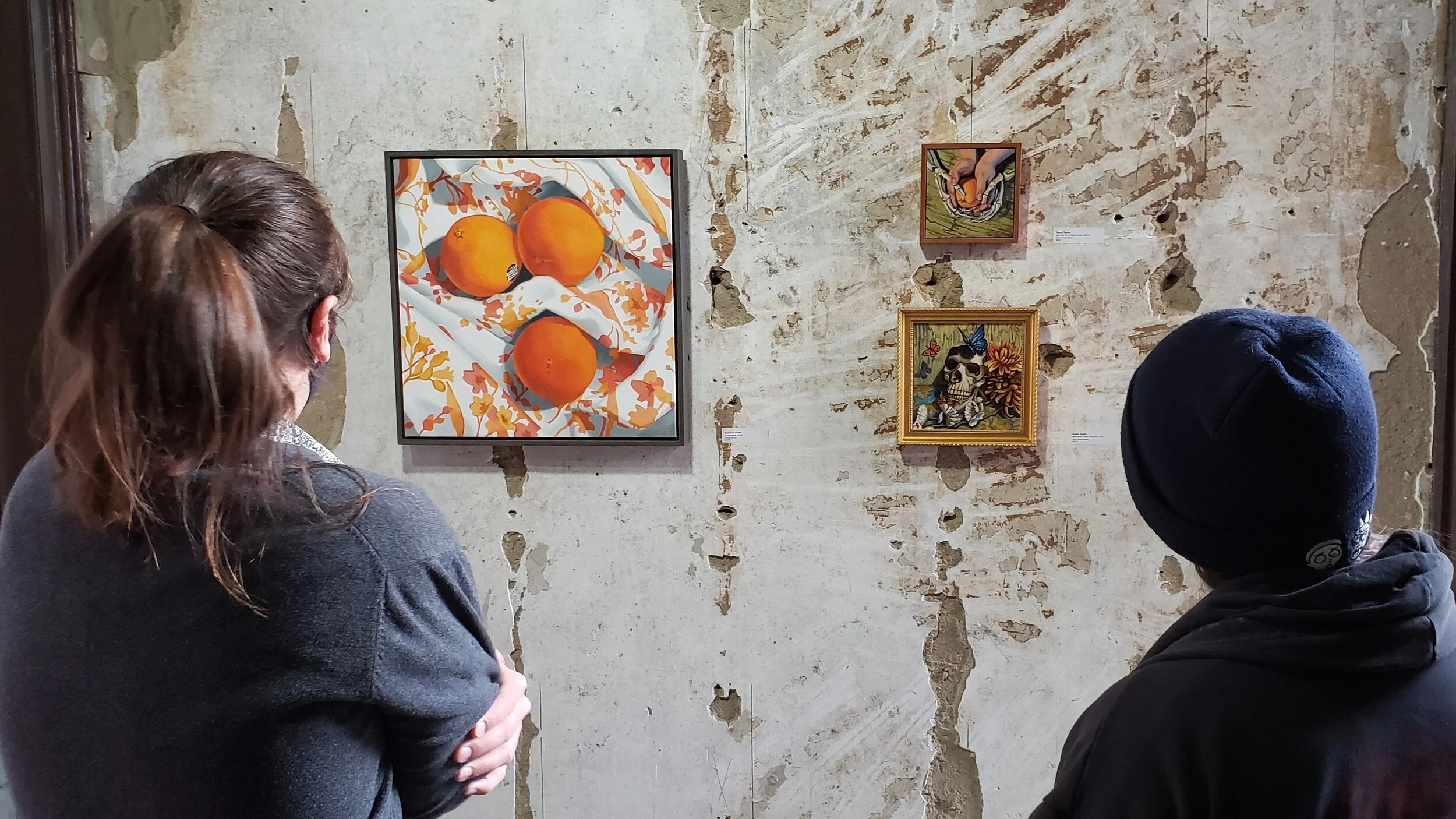 Two people viewing artwork on a textured wall with peeling paint in an art gallery. The artwork includes a painting of three oranges on a patterned cloth, a small piece with hands holding a fruit, and two framed images of a skull with butterflies and