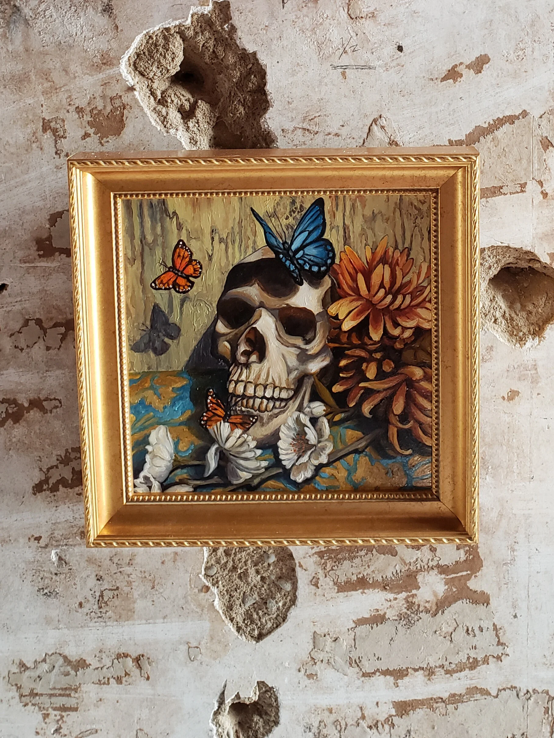 Painting of a human skull surrounded by colorful butterflies, flowers, and a wood-like background, framed in a gold ornate frame, mounted on a distressed brick wall.