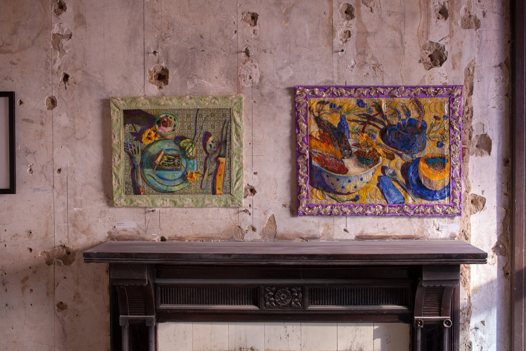 Two colorful embroidered tapestries hanging on a deteriorated wall above a dark wooden fireplace mantel.
