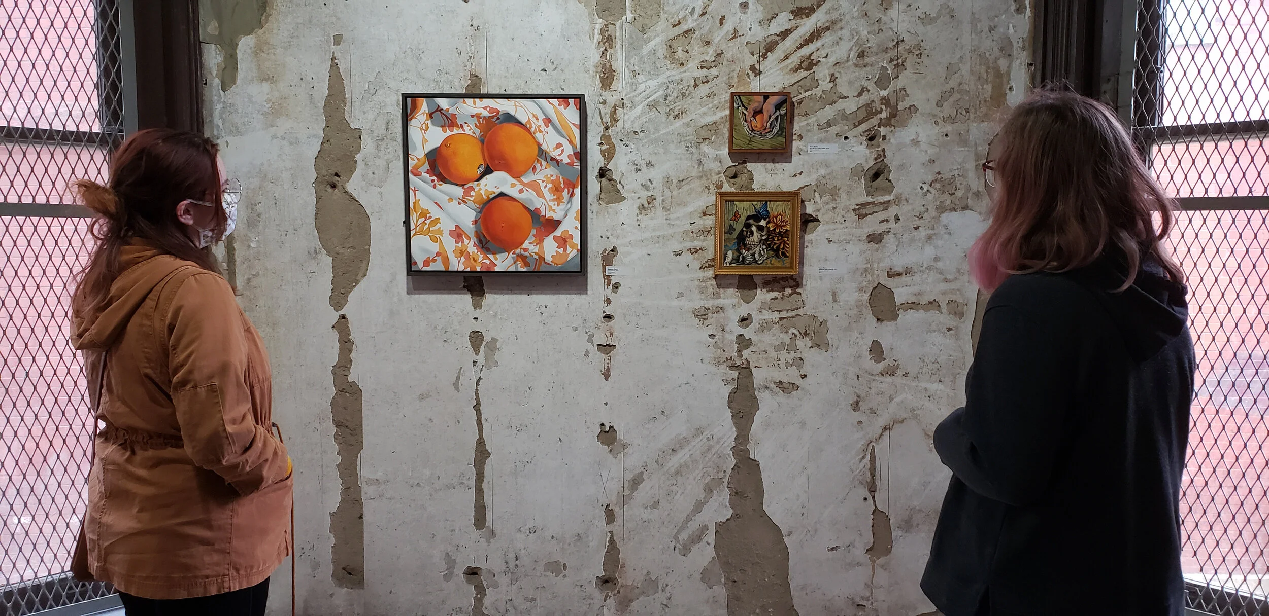 Two women observing artwork on a distressed concrete wall in an art gallery. One woman is wearing a brown jacket and a face mask, the other is wearing a dark jacket with pink hair.