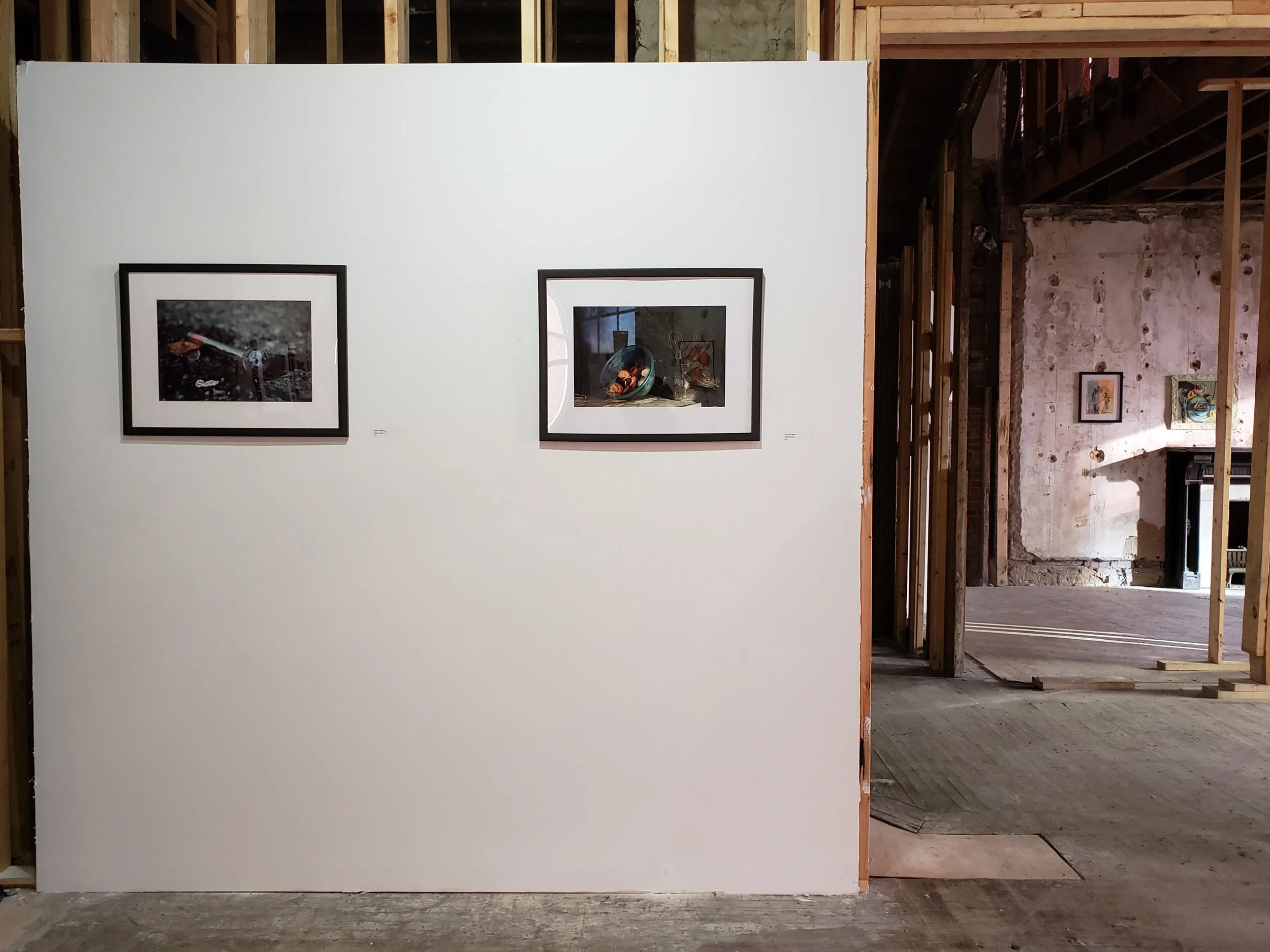 Two framed photographs hung side by side on a white wall in an art gallery or museum, with an unfinished interior of a building visible in the background.