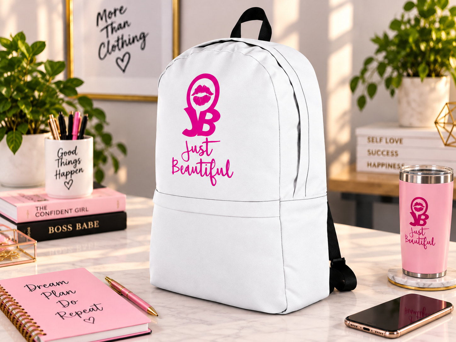 A white backpack with pink and black lettering that says "JB Just Beautiful" and a pink lip print, placed on a white marble table surrounded by notebooks, a pen, a tumbler, and decorative items.