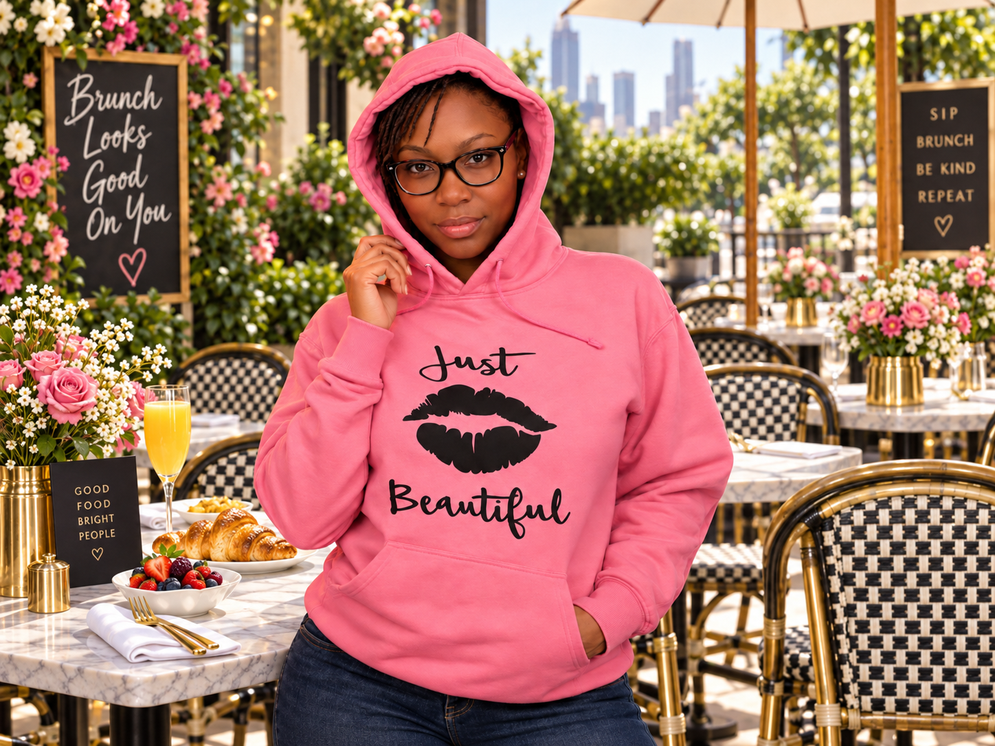 Young woman wearing a pink hoodie with a lips graphic and 'Just Beautiful' written on it, standing in an outdoor brunch setting decorated with pink and white flowers, with a city skyline in the background.