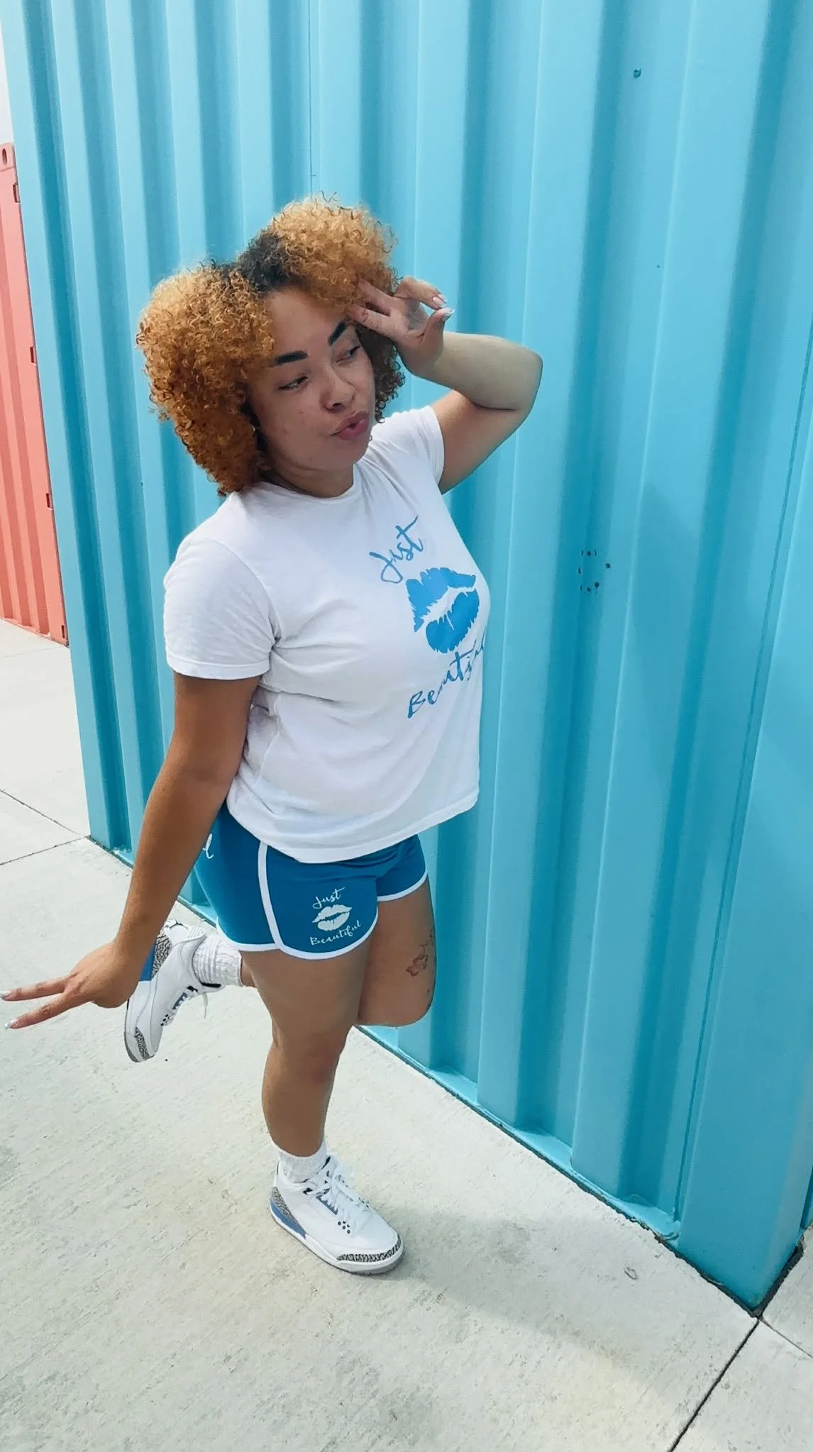 A woman striking a playful pose in front of blue lockers, wearing a white t-shirt with a blue lip print and the words 'Just Beautiful', blue shorts with similar print, and white sneakers.