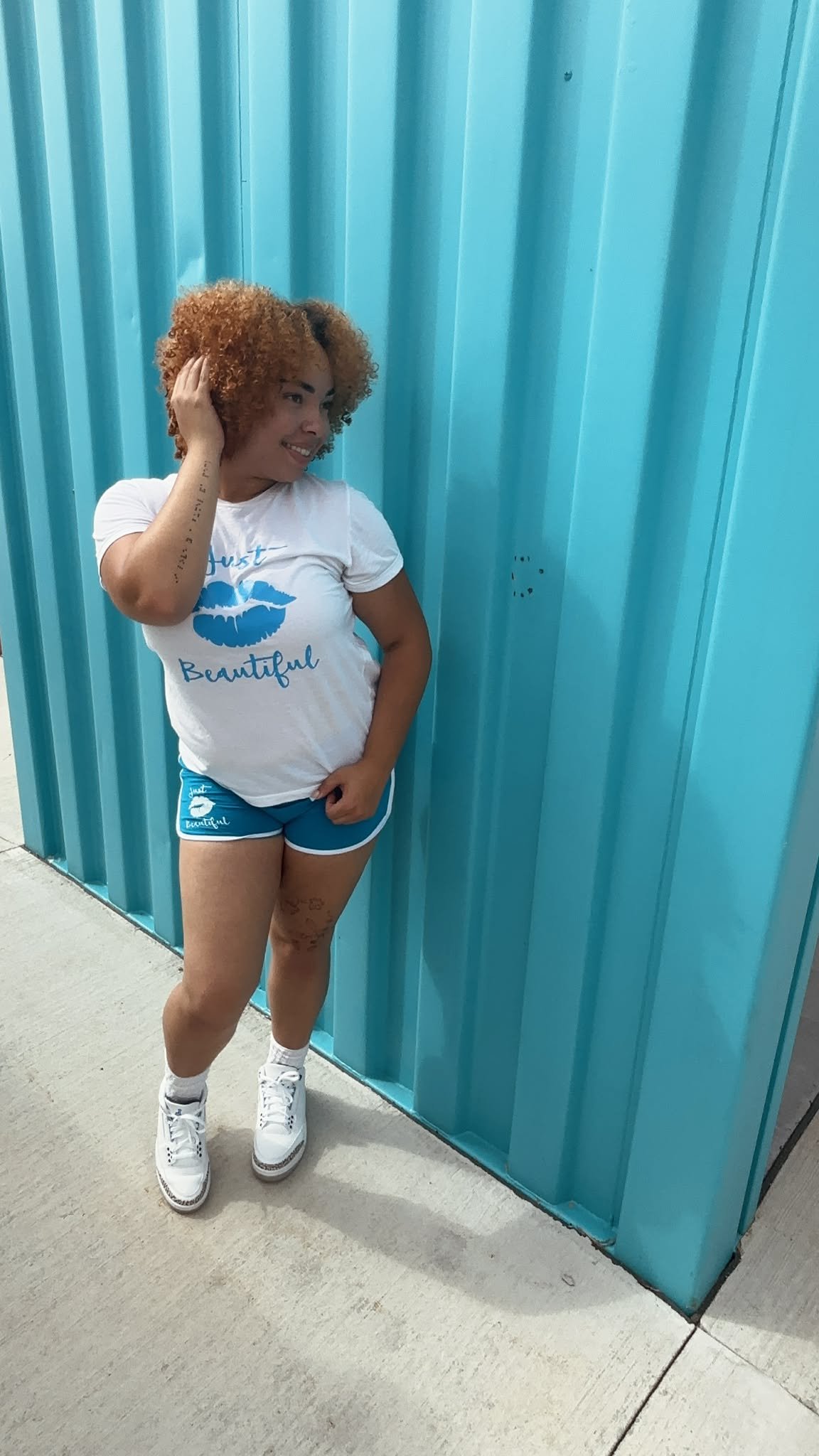 A woman with curly hair standing against a blue corrugated metal wall. She is wearing a white T-shirt with a blue graphic of lips and the words 'Just Beautiful,' matching blue shorts, white sneakers, and white socks. She is smiling and touching her h