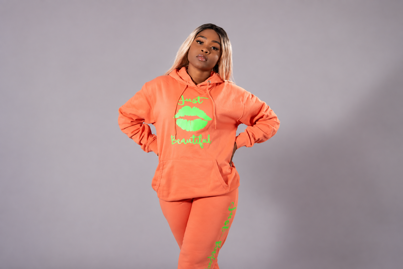 A young woman in an orange hoodie and sweatpants with neon green text and graphics posing with hands on hips against a gray background.