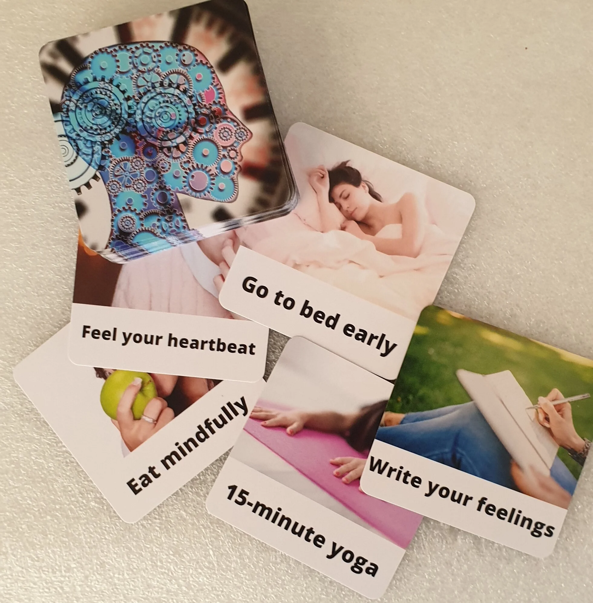 A collection of wellness and self-care cards including a card with mechanical gears forming a human head profile, a woman sleeping, a person doing yoga, someone writing feelings, a hand holding an apple, and a note to go to bed early. Each card has different instructions or prompts.