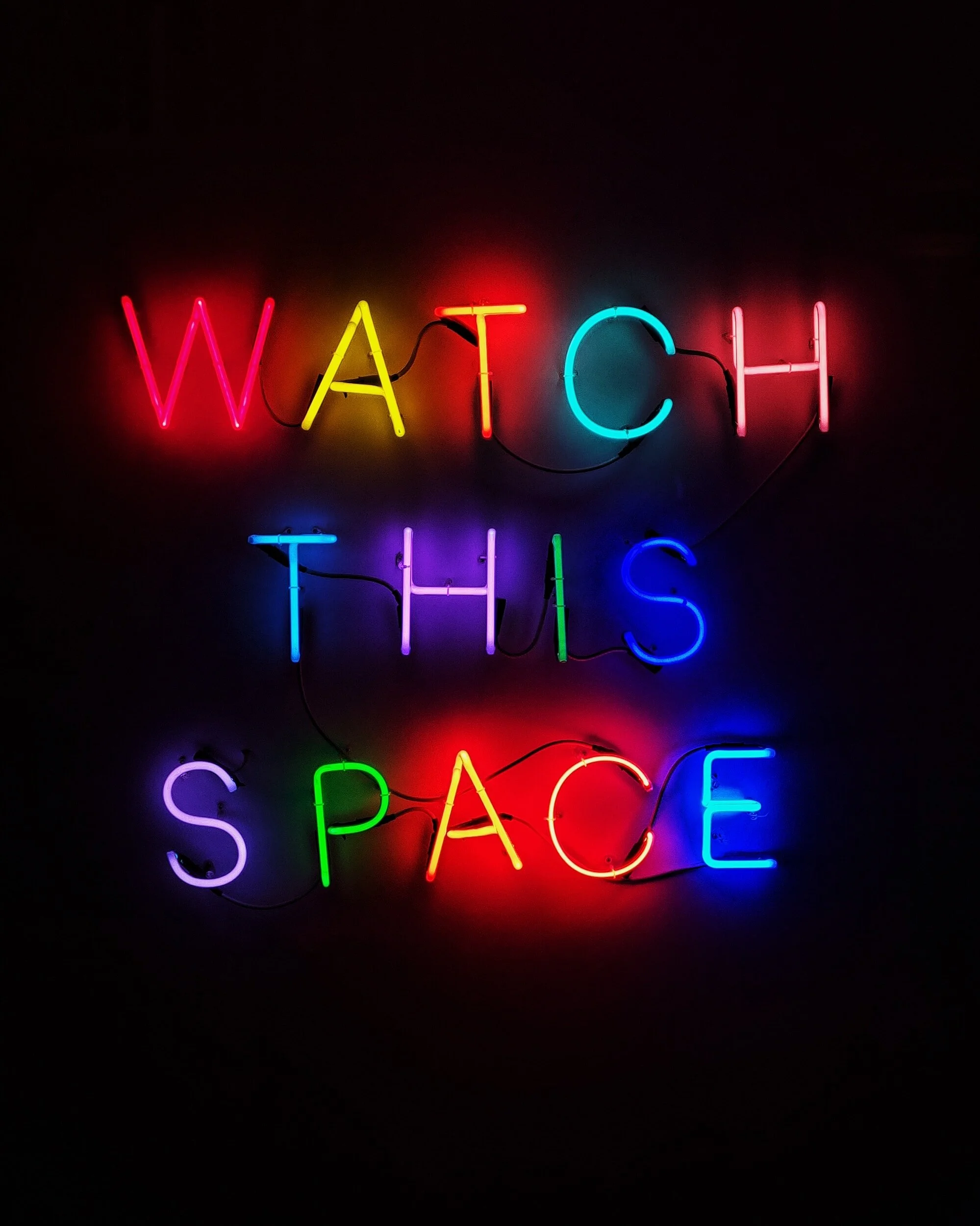 Neon sign with colorful letters spelling 'WATCH THIS SPACE' against a dark background.