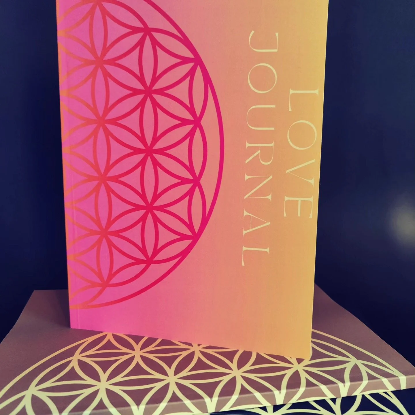 A pink love note booklet with a geometric flower of life pattern on the cover, placed on top of a similar patterned paper. The words ‘Love YourN’ are written vertically on the right side of the booklet.