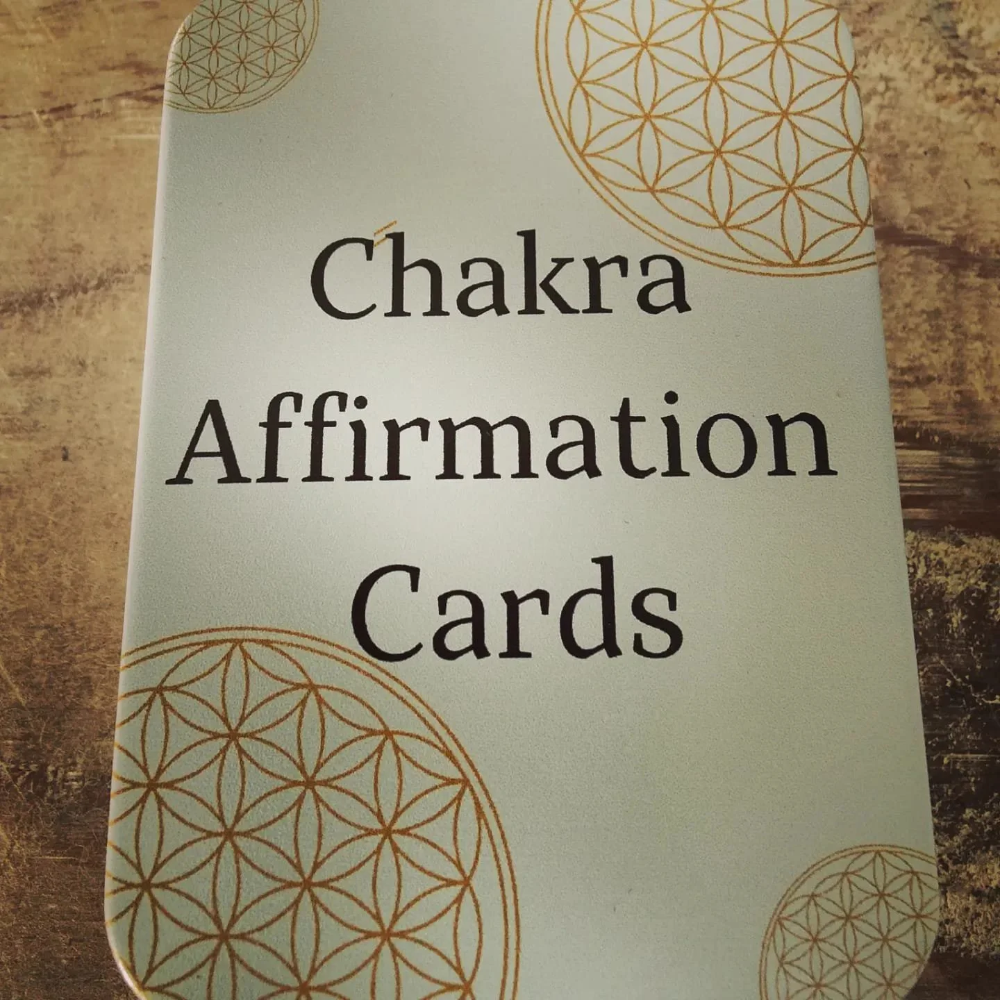 A pack of Chakra Affirmation Cards with a light-colored background and geometric designs in gold.