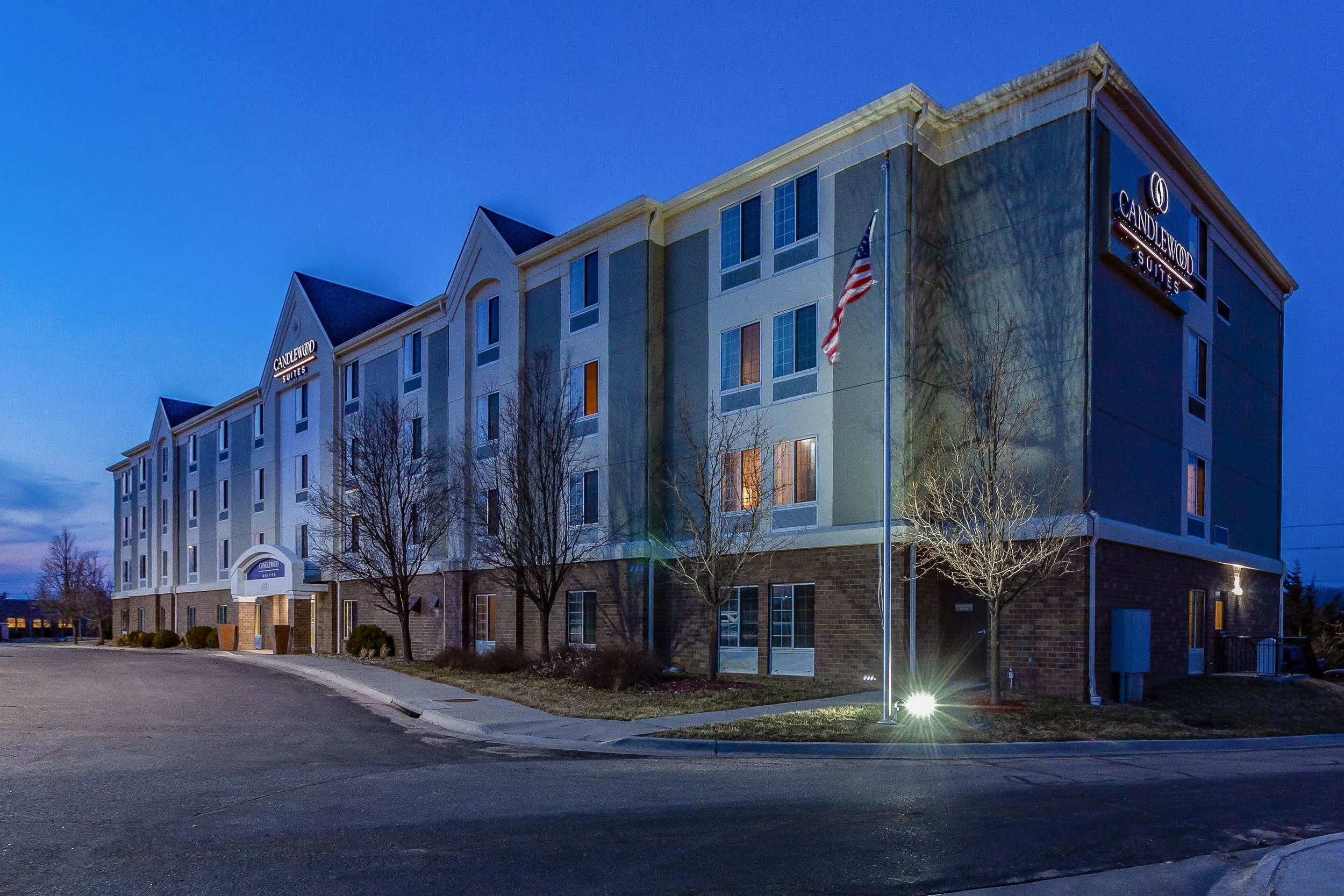 Nighttime exterior view of Candlewood Suites hotel with an American flag, trees, and a sidewalk in front.