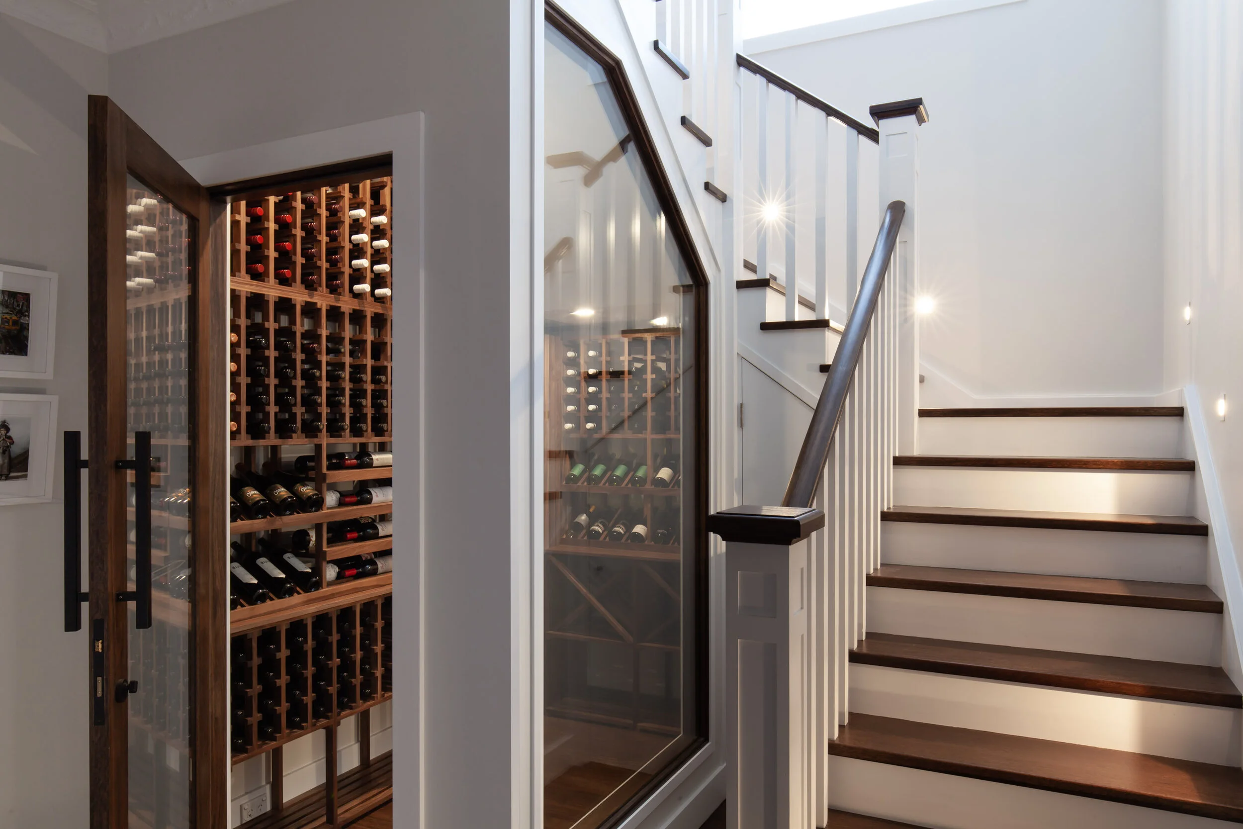 First floor addition with a wine celler under the stairs. A staircase with wooden steps and white railings next to a wine cellar with wine bottles stored in wooden racks behind glass doors.
