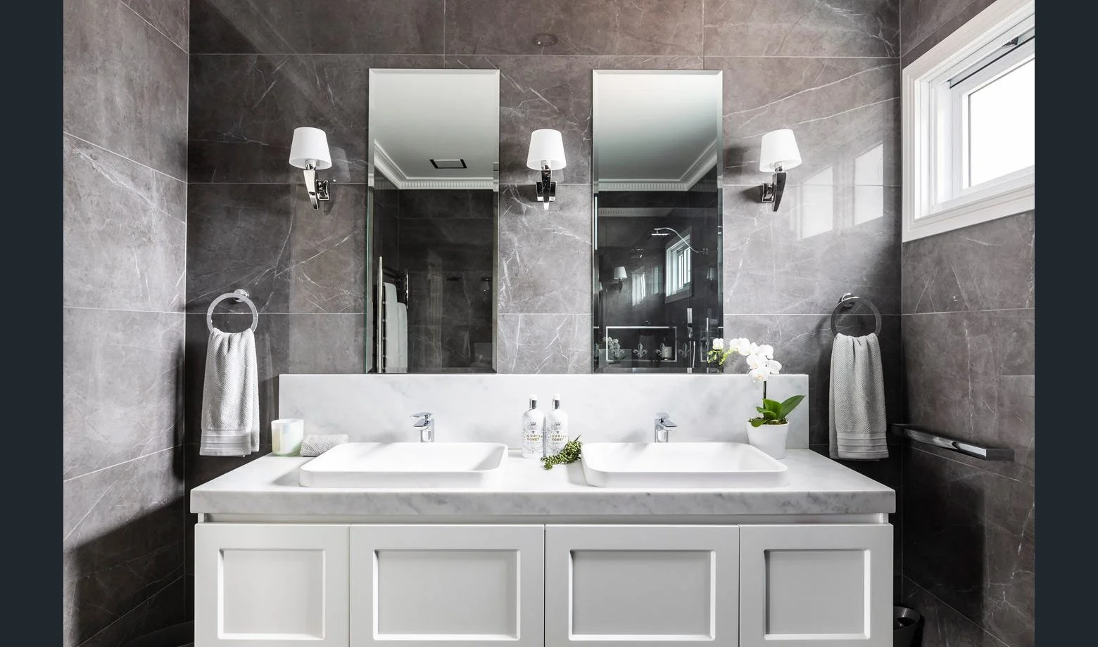 Second storey modern bathroom with twin white sinks and large mirror, brown marble walls, two wall-mounted light fixtures, a window on the right.
