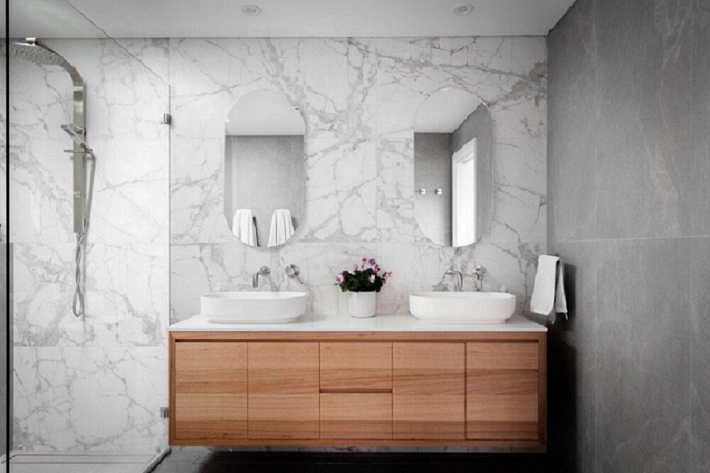 Second storey addition modern bathroom with double vanity, oval mirrors, marble wall, wooden cabinet, potted plant, and walk-in shower with glass door.