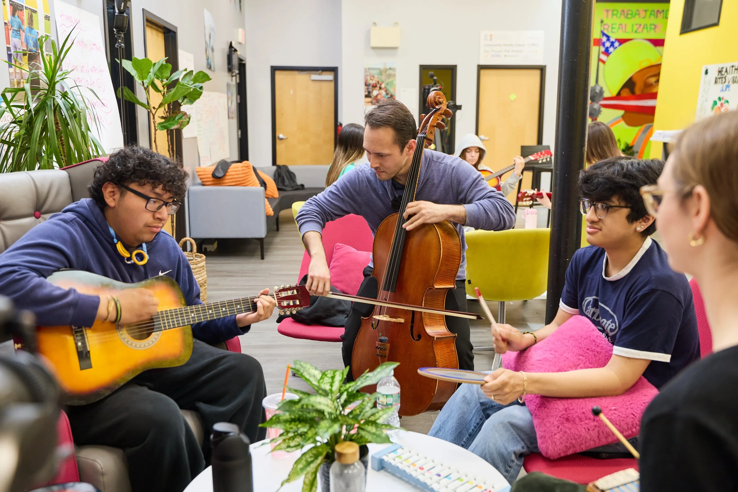 A cellist plays alongside young musicians on guitar and percussion during a collaborative music workshop in a colorful community space.