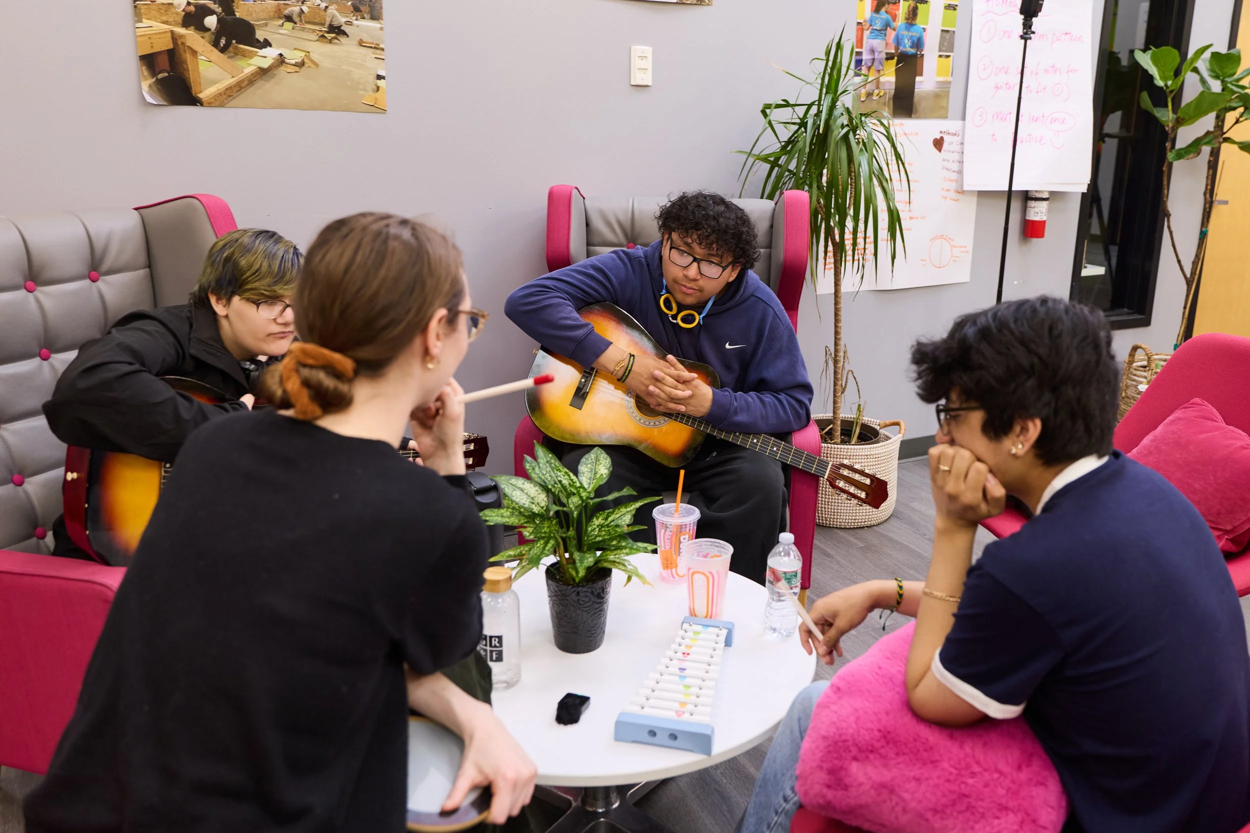Young musicians gather in a community space with acoustic guitars and percussion instruments, listening and sharing ideas during a collaborative music workshop.