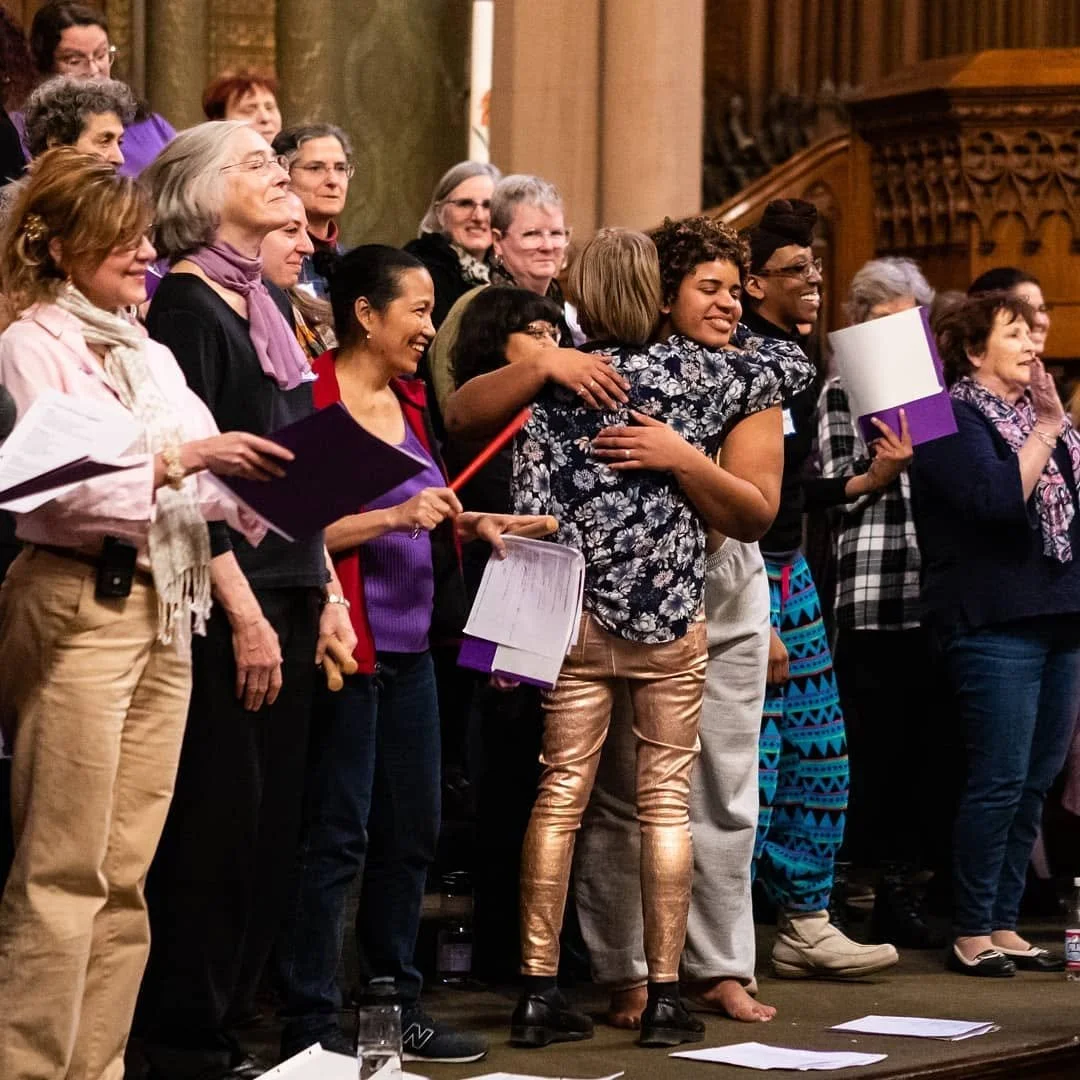 A diverse group of women choir members smiling, embracing, and holding sheet music during a choral performance inside a church.