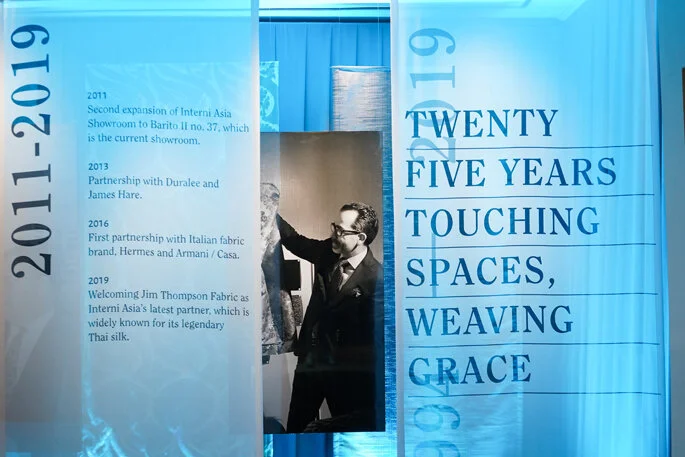 Display wall with timeline of events from 2011 to 2019 related to Interni Asia, featuring a man in a suit looking at fabric samples, with large blue text reading 'Twenty Five Years Touching Spaces, Weaving Grace'.