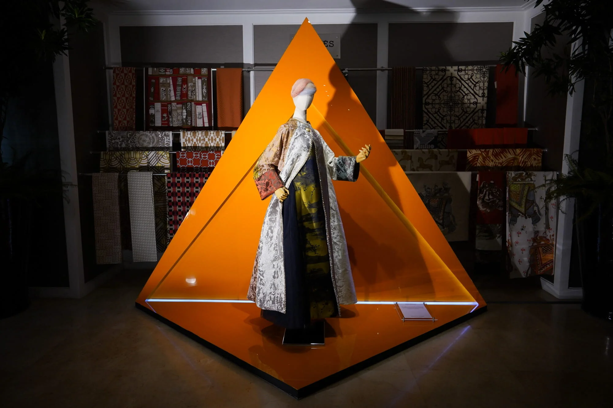 A mannequin dressed in an ornate, traditional garment displayed inside an orange, pyramid-shaped enclosure with bright lighting, against a background of shelves with fabric samples.