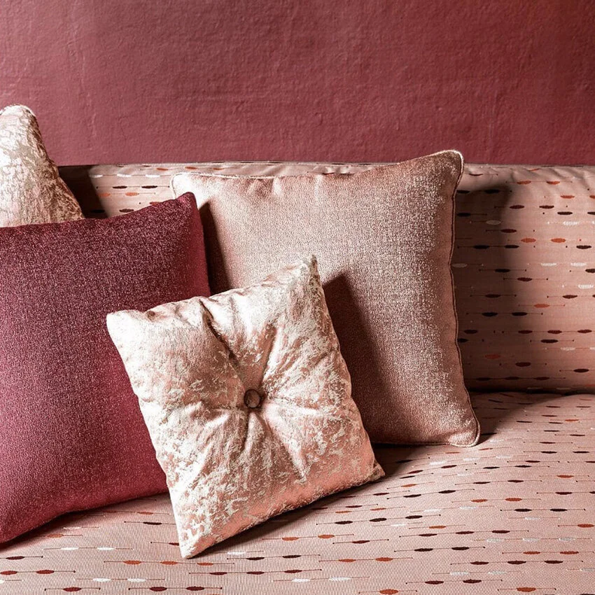 A collection of pink and velvet cushions on a pink wall and patterned pink sofa.