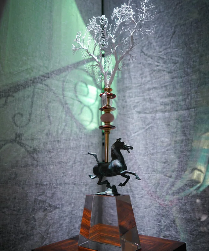 An ornate sculpture featuring a black horse with raised front legs, mounted on a reflective metallic base, with a decorative pole topped with a tree-shaped shadow projecting onto the background.