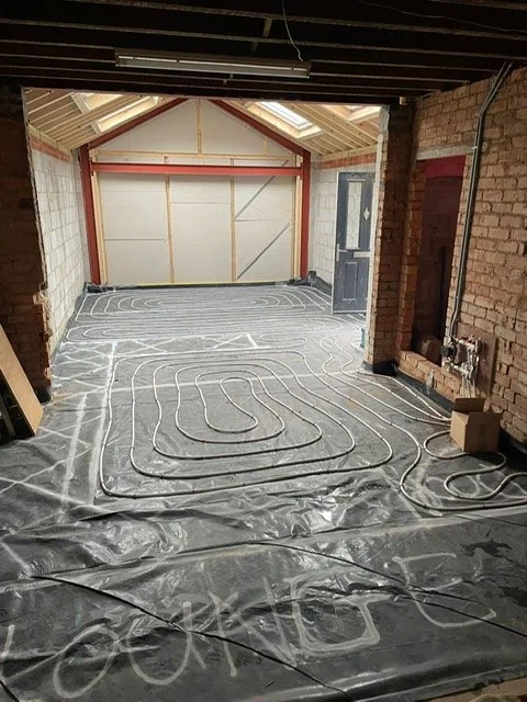 Room under renovation with underfloor heating coils installed on a concrete floor, exposed brick walls, and partial ceiling beams.