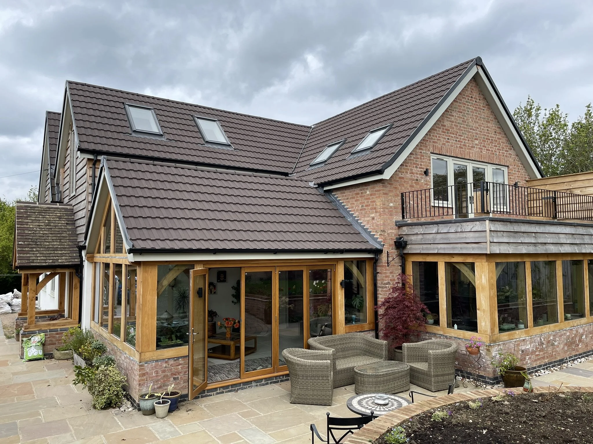 Modern brick house with brown roof, large windows, patio furniture, and plants.