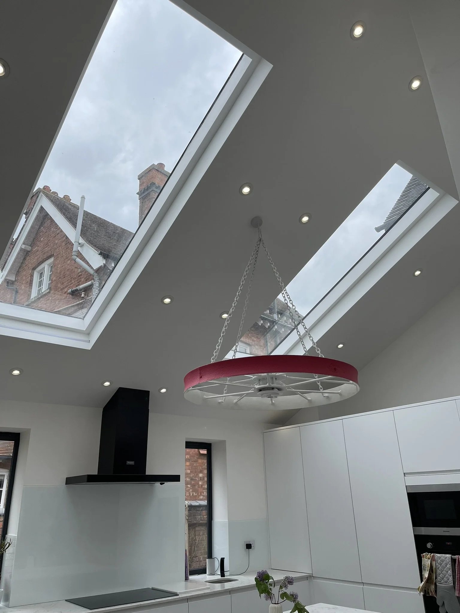 Modern kitchen with large skylights, suspended decorative wheel, ceiling lights, white cabinetry, and black range hood.