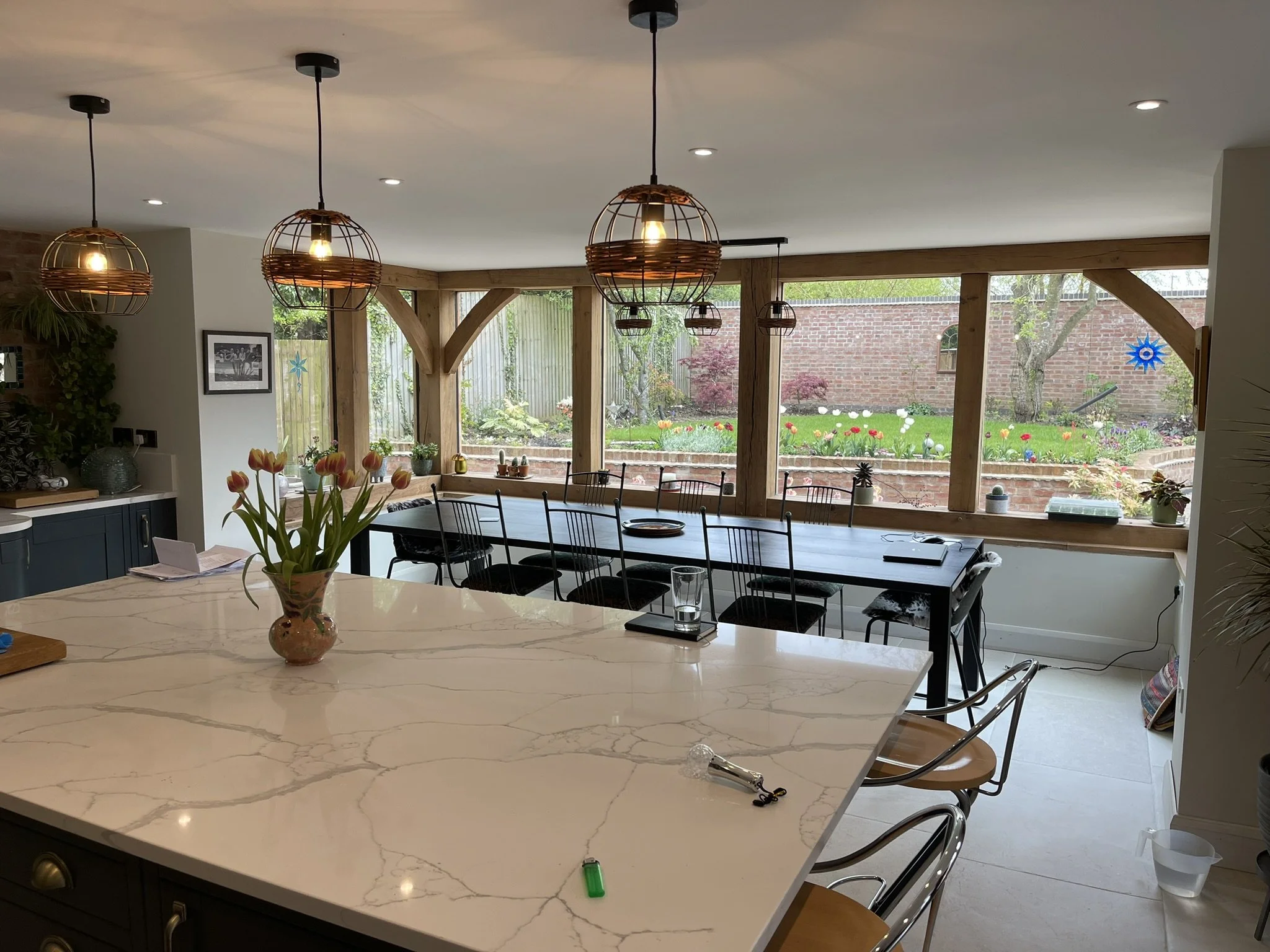 Modern kitchen with large marble countertop, pendant lights, potted flowers, and glass wall overlooking a garden.