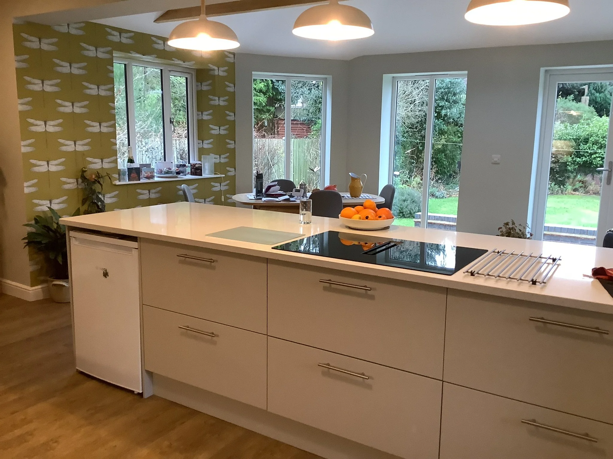 Modern kitchen with island, induction cooktop, white cabinets, and dragonfly-patterned wallpaper. Three pendant lights hang above. Large windows and glass doors provide a view to a green garden. 