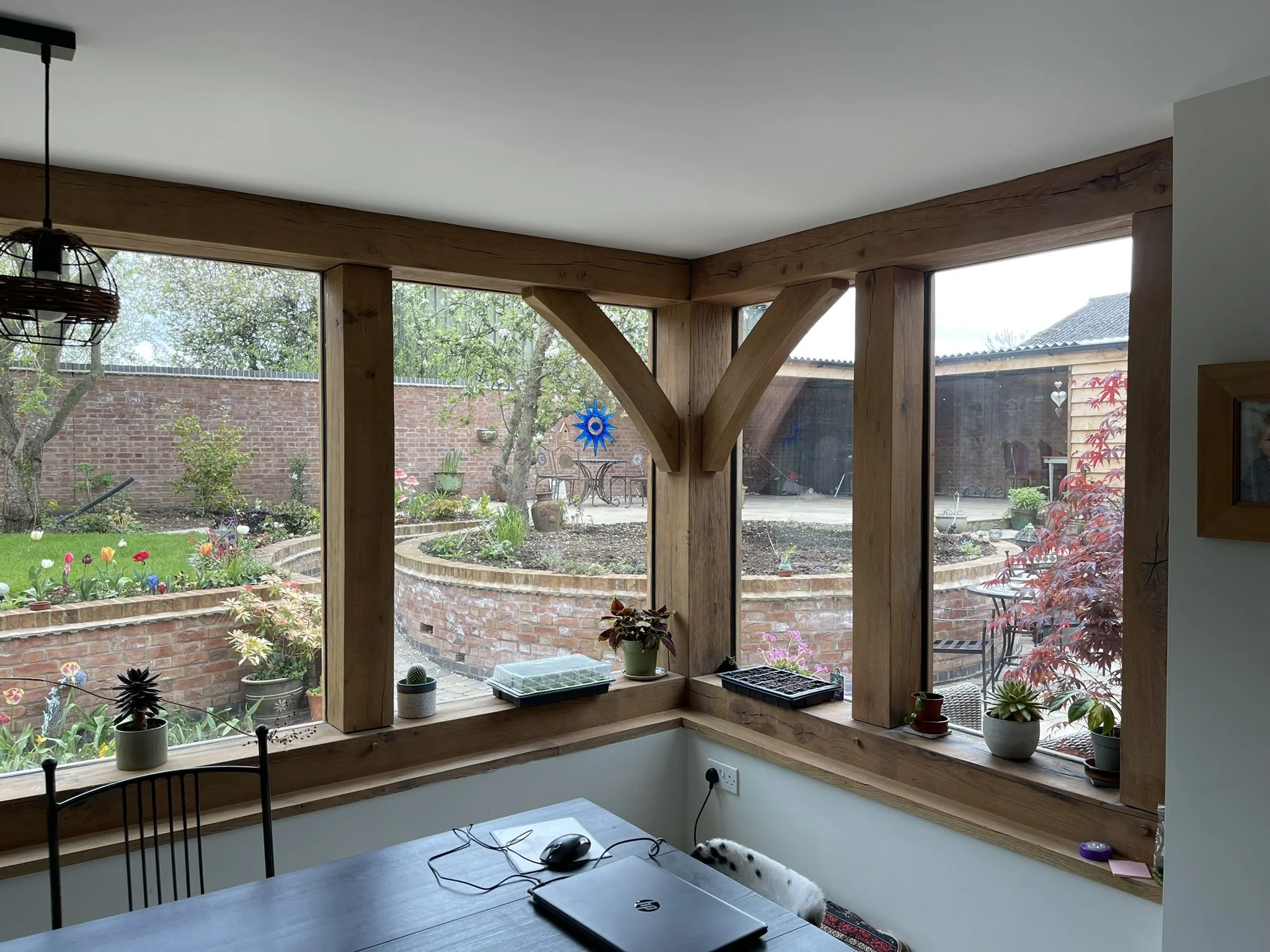 Interior view of a room with wooden beams, overlooking a garden with a brick wall, grass, flowers, and trees. Small potted plants are on the windowsill, and a laptop and mouse are on the table inside.