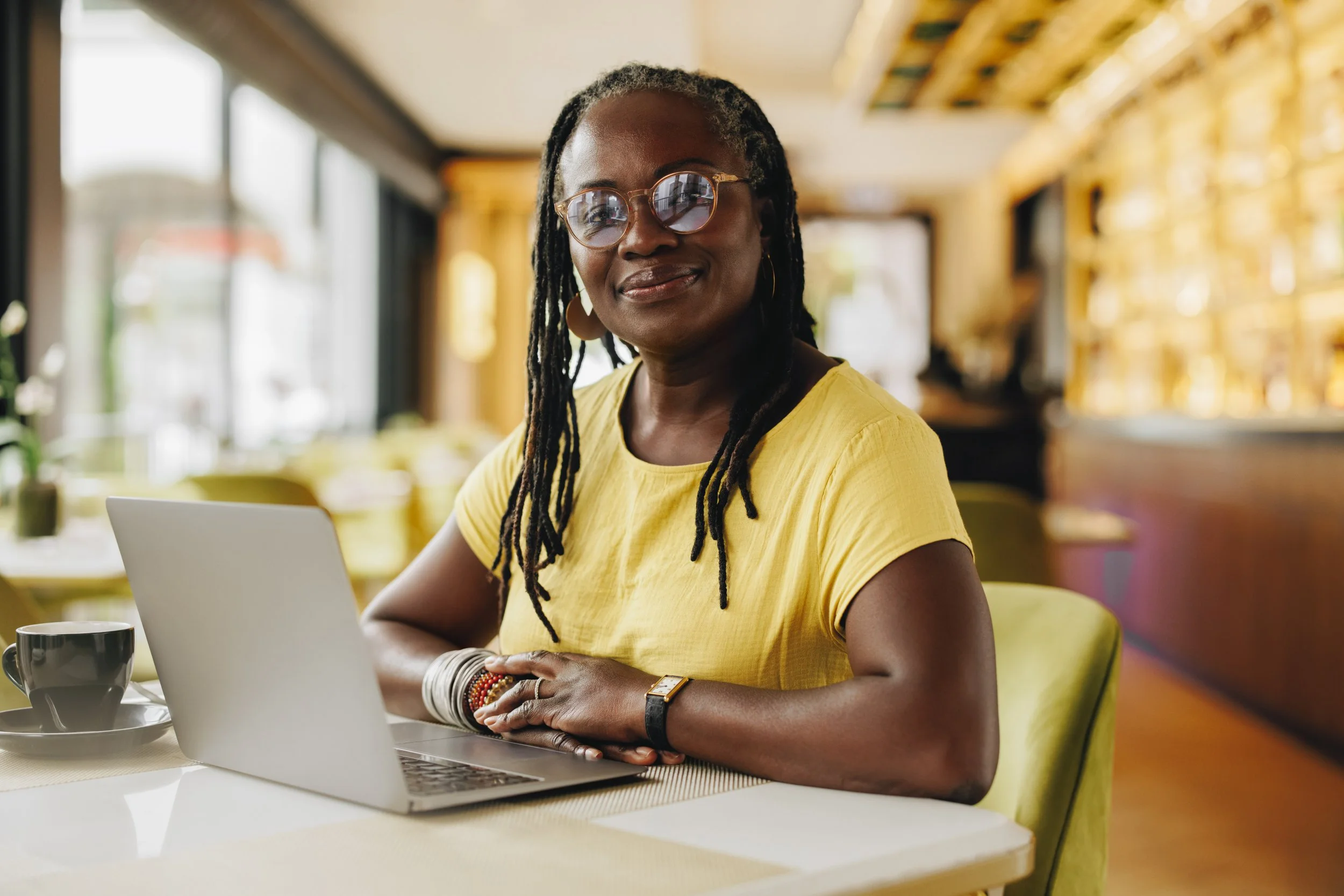 A woman with glasses and dreadlocks sitting at a table in a cafe, smiling, wearing a yellow shirt, with a laptop in front of her and a cup beside it.