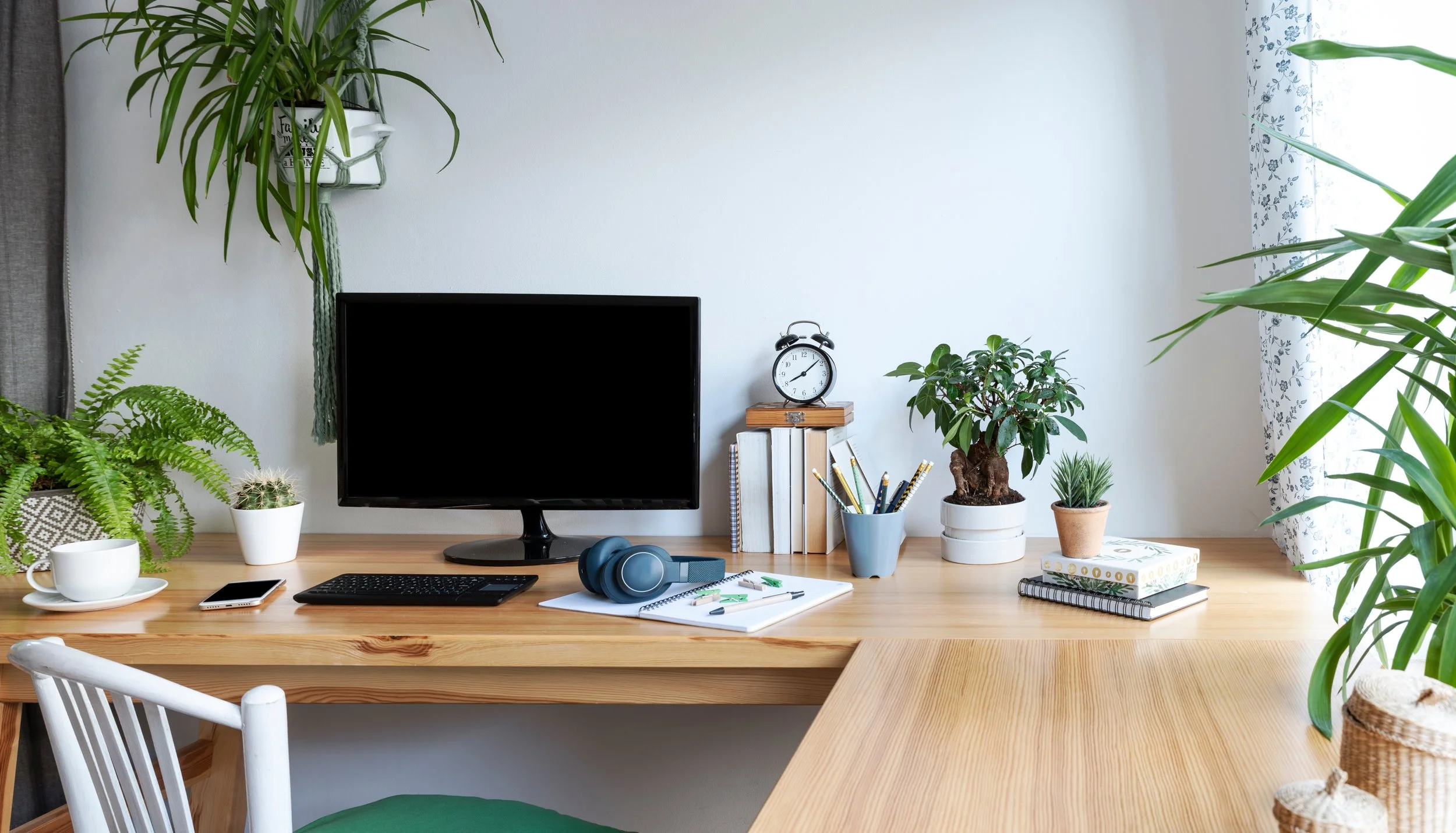 A wooden desk with various items including a computer monitor, keyboard, headphones, notepads, pens, a smartphone, a cup and saucer, and several potted plants in a room with white walls and floral curtains.