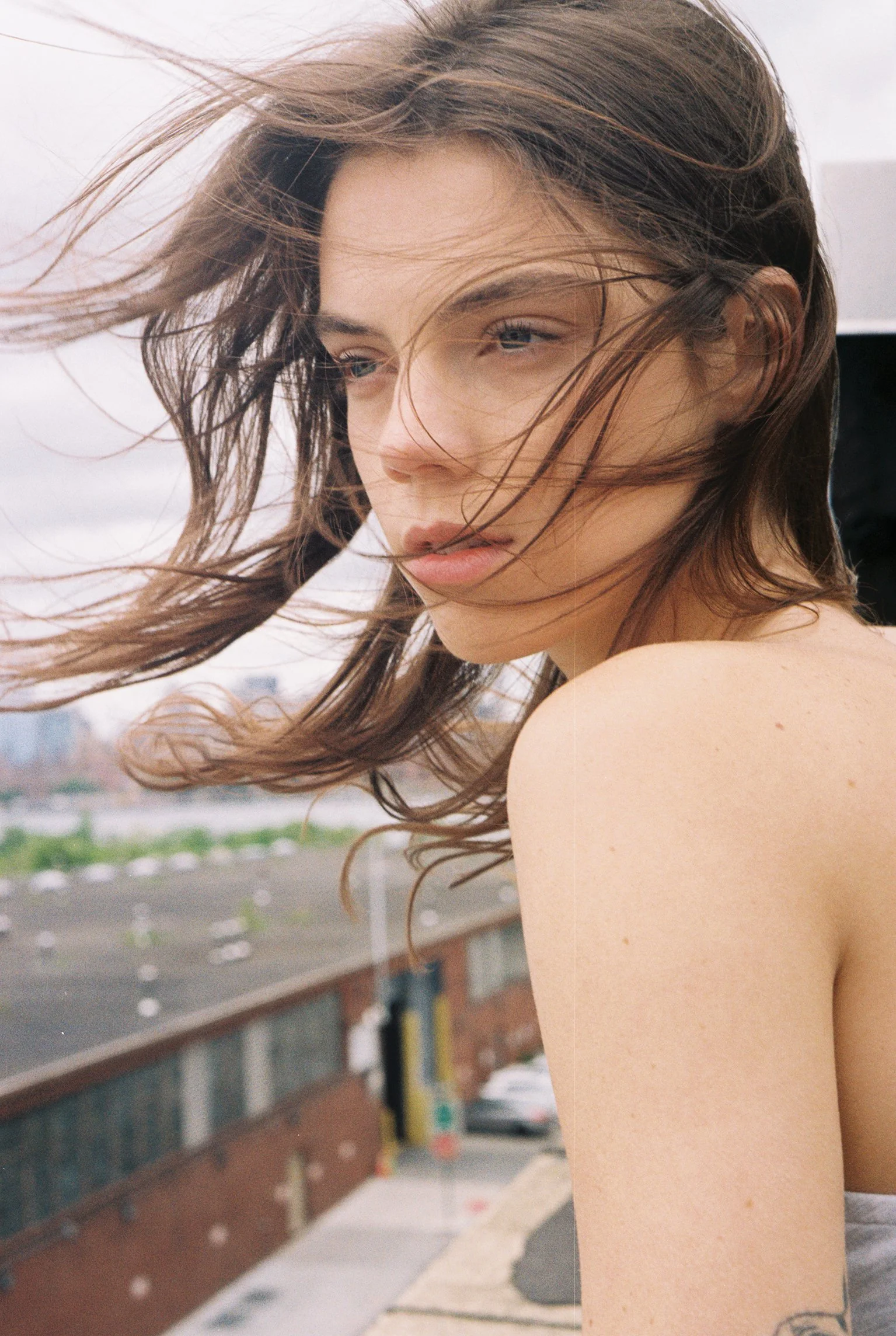 A woman with long, flowing brown hair outdoors with a blurred background of a building and a parking lot.