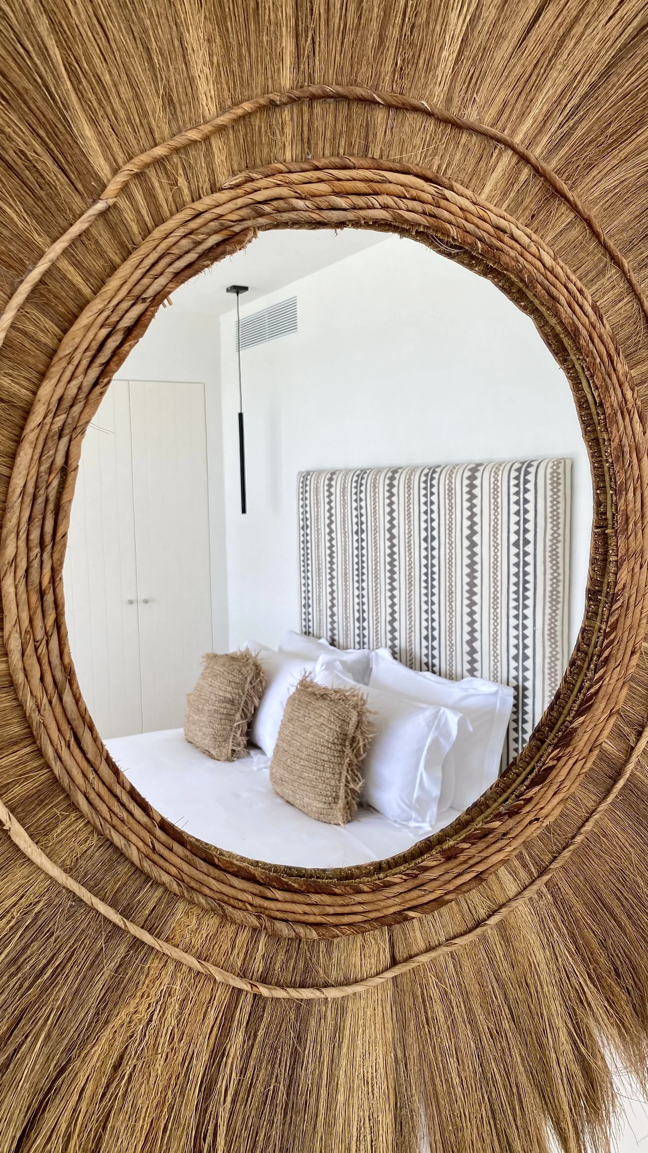Reflection of a bedroom in a circular woven mirror showing a bed with a striped headboard, white bedding, and two beige textured pillows.