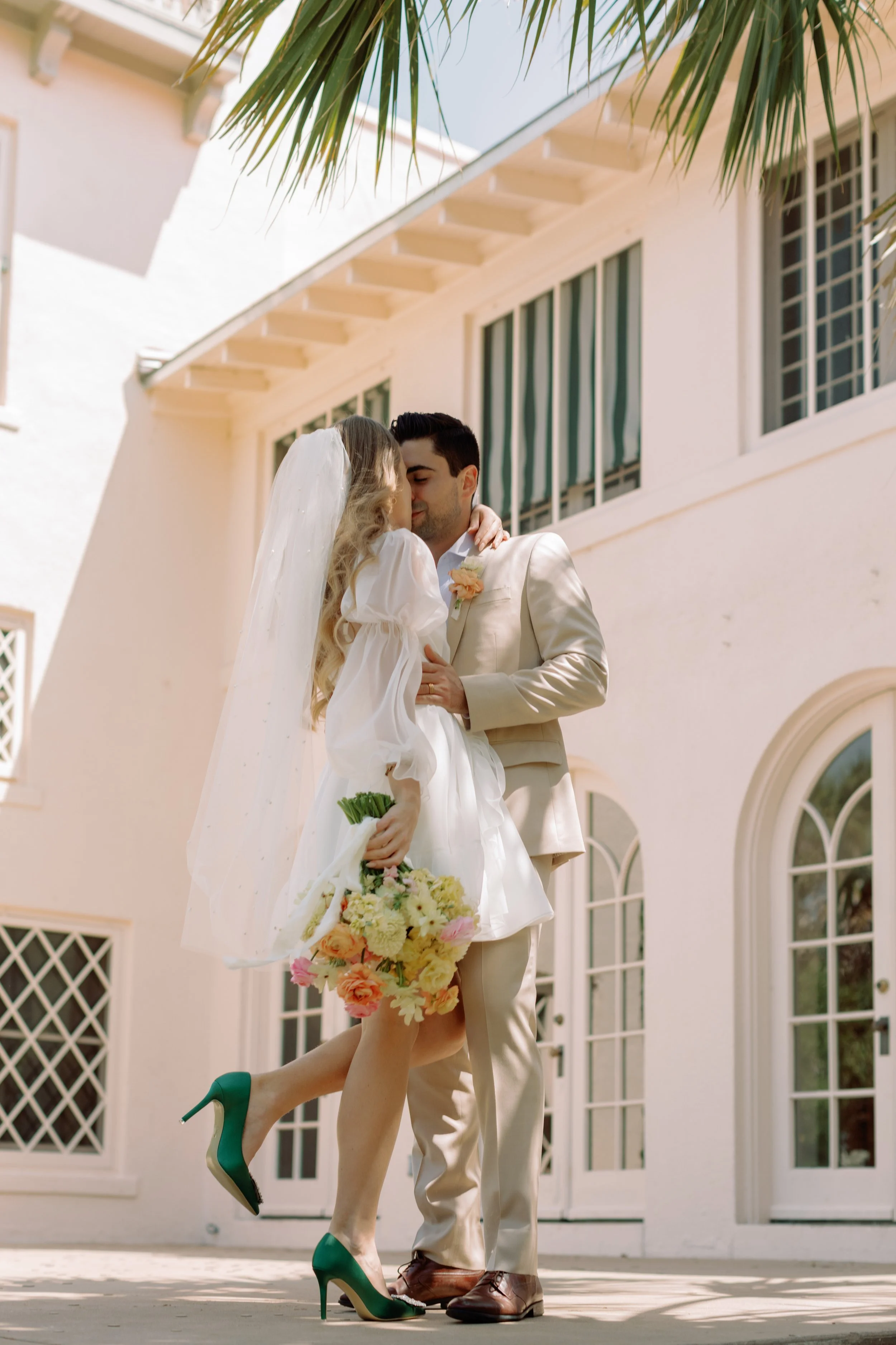 couple in front of a large building standing outside together in the palm trees, in the style of light emerald and light beige, vintage elegance, vibrant florals, dynamic pose, light orange and gold, light-filled,