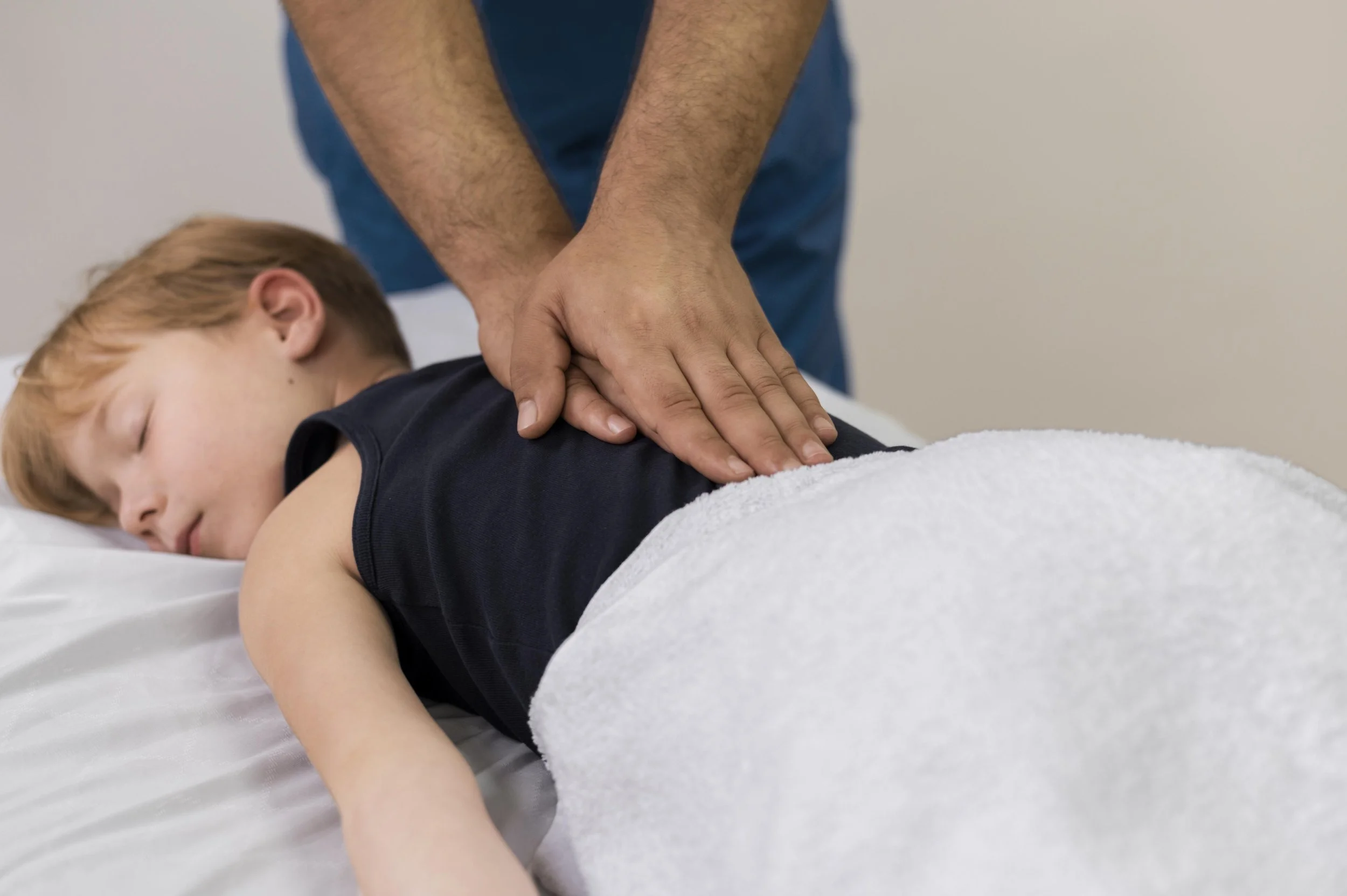 A person performing CPR on a sleeping child lying face down on a bed, with a towel partially covering the child, in a medical setting.